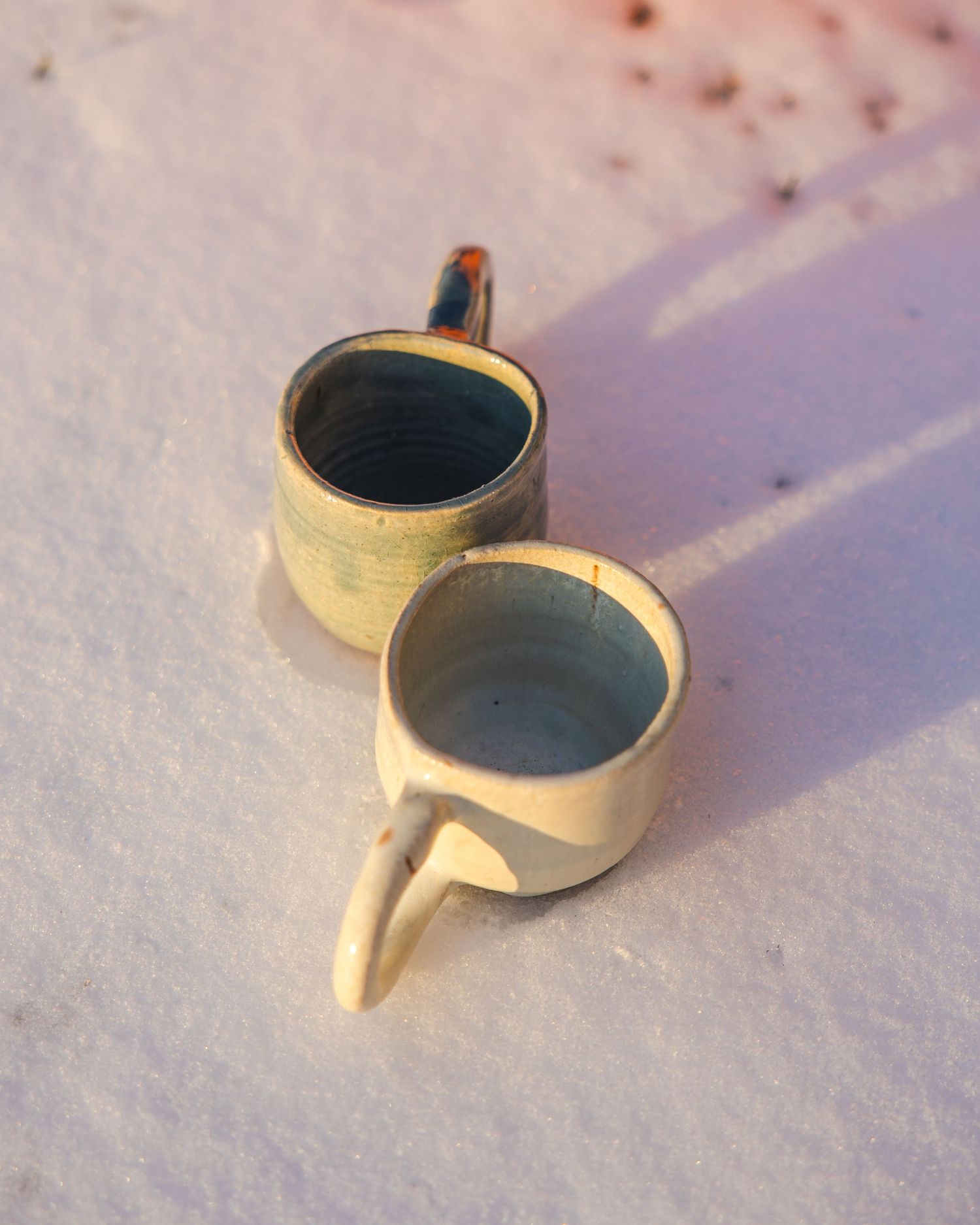 Two handmade ceramic mugs with teal glaze interior resting in white snow.