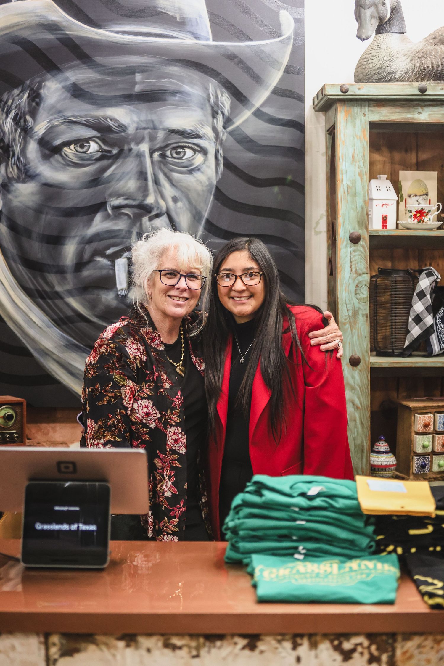 Two smiling women pose together at a Grasslands of Texas retail shop counter with western mural backdrop.