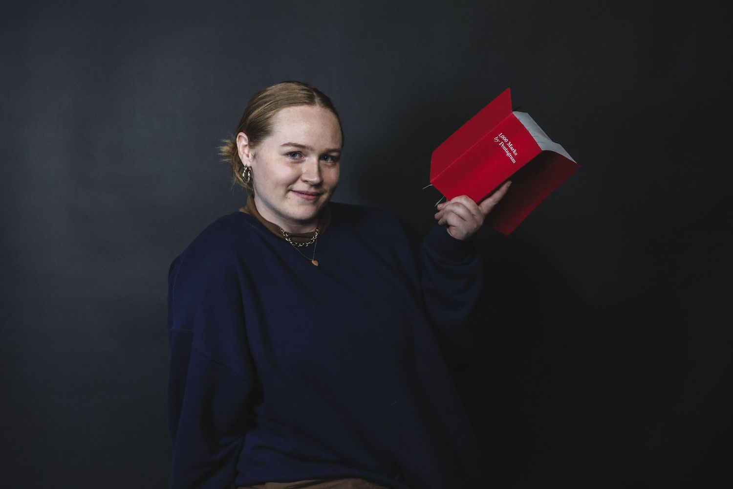 Smiling young woman in navy sweatshirt holding red book against dark background.
