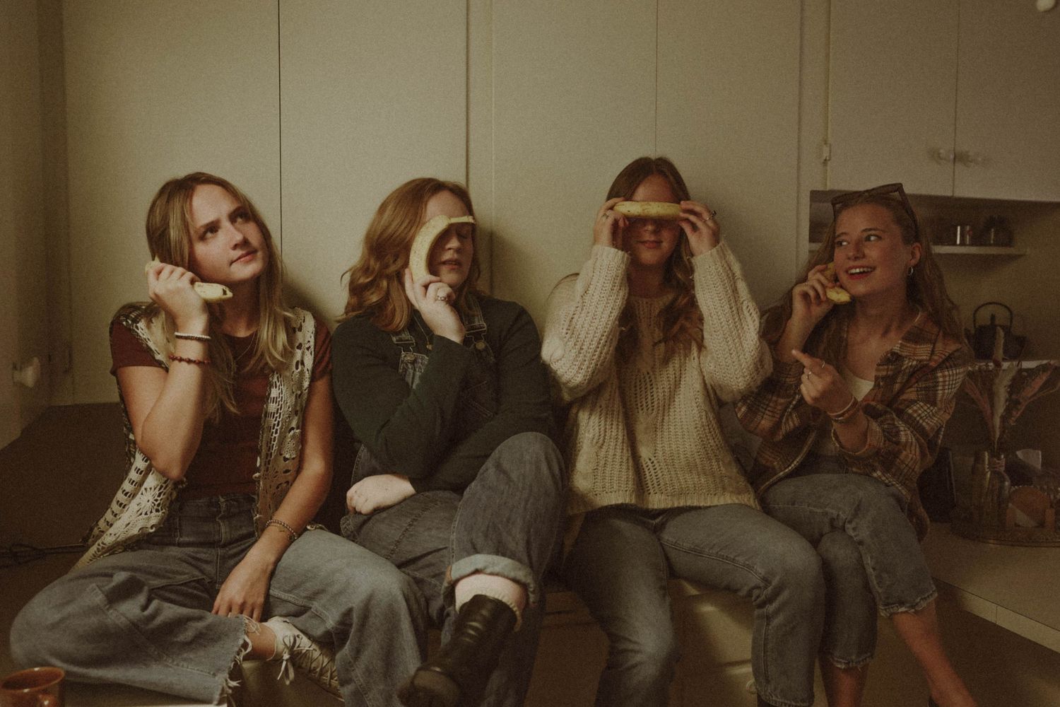 Four young women sitting together playfully covering eyes with hands and banana slices indoors.