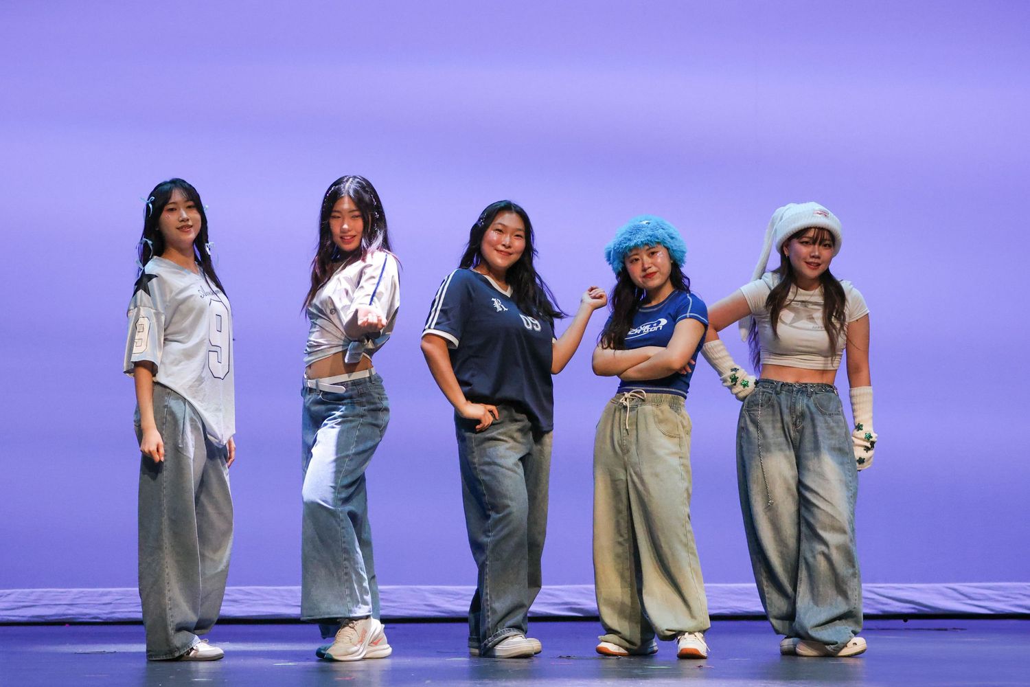 Five young women posing confidently on stage in casual streetwear against a purple backdrop.