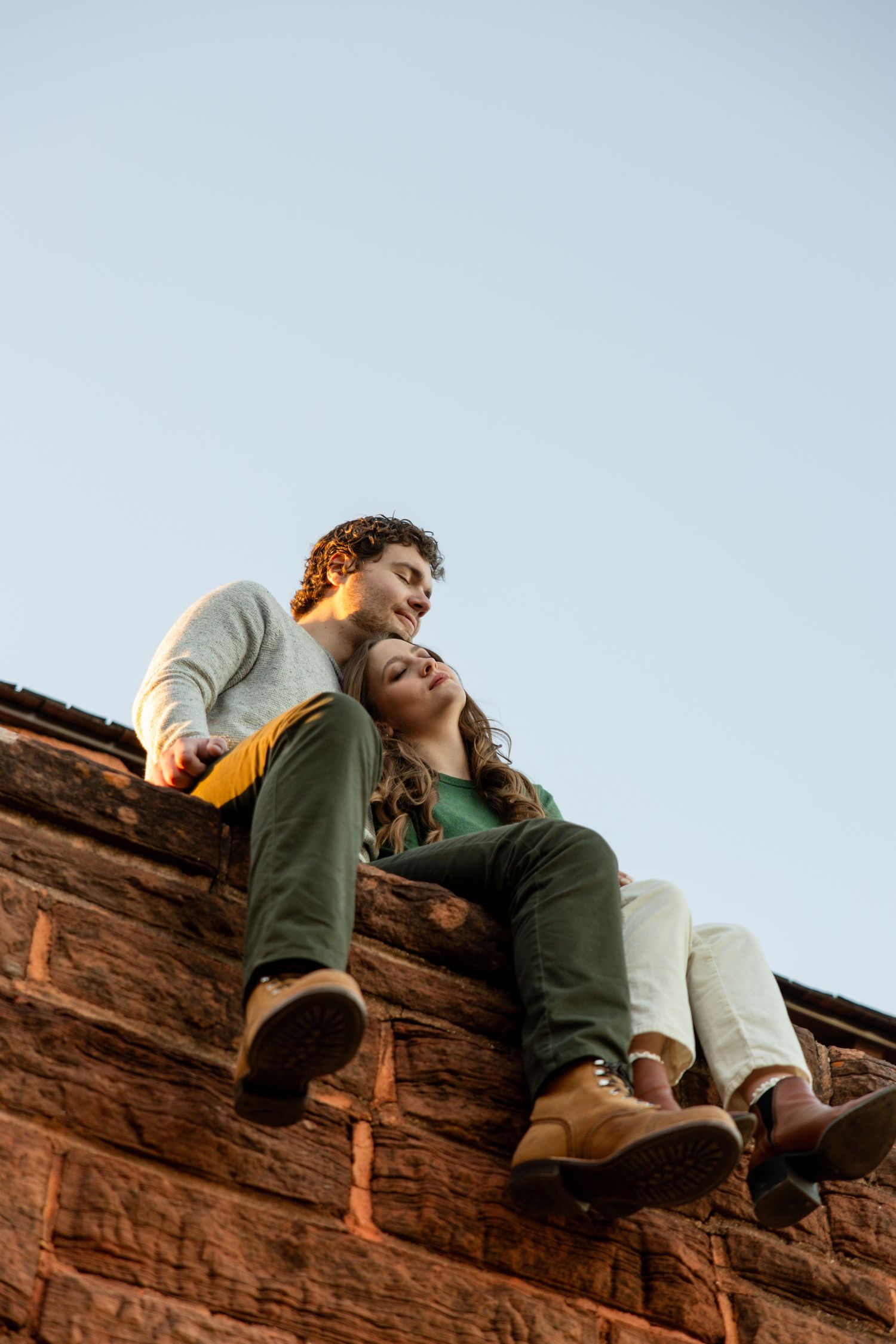 Couple sitting on top of a rustic brick wall at dusk, leaning together against a clear blue sky.
