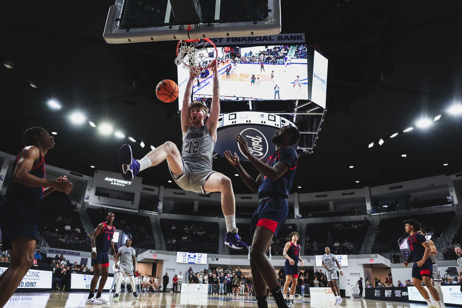 A gray-uniformed player completes a slam dunk as red-uniformed defenders watch during a college basketball game.