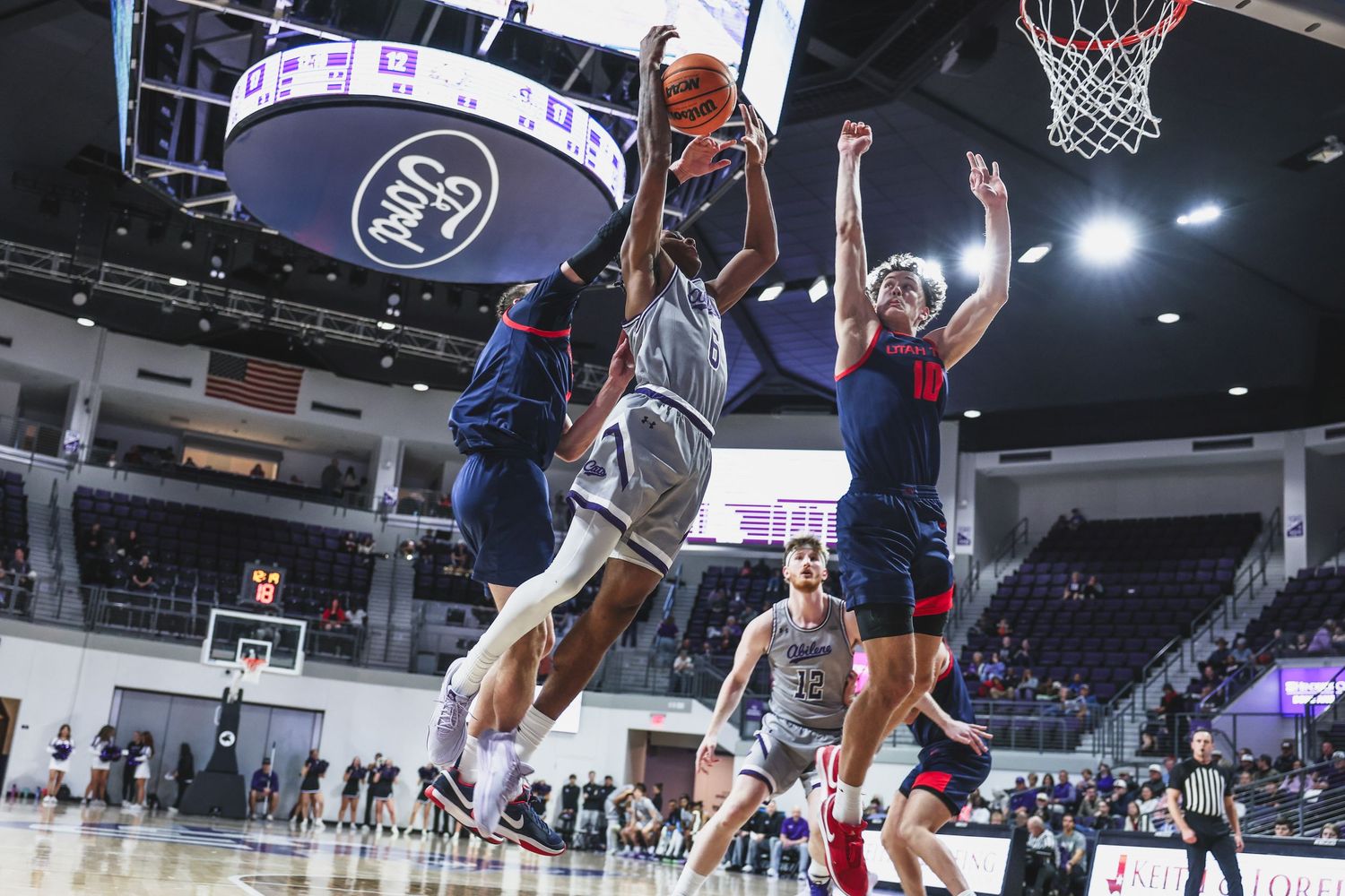 College basketball player drives between two defenders for a layup in a packed indoor arena.