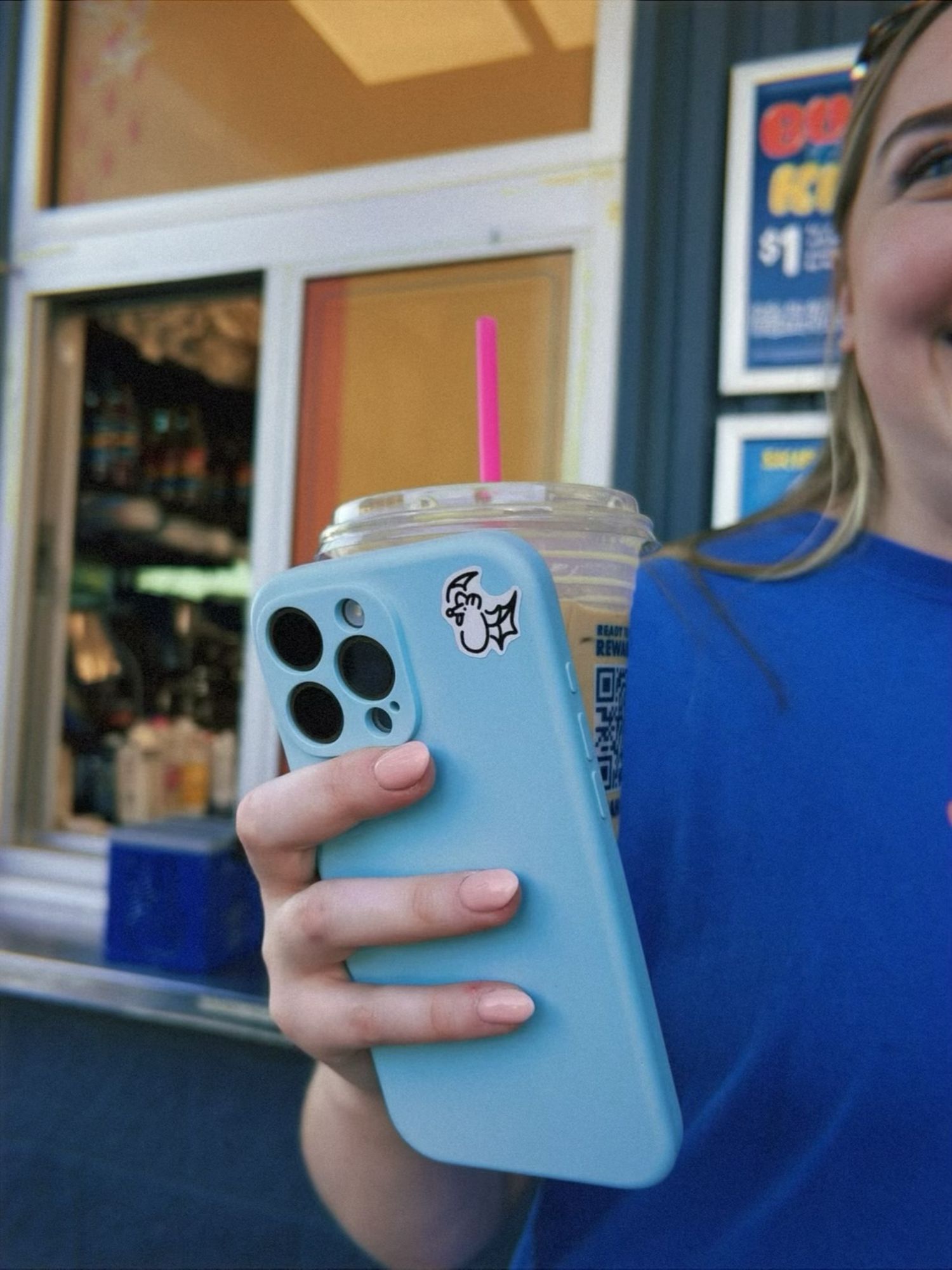 Girl holding iced coffee drink with pink straw and light blue iPhone case outside a shop.