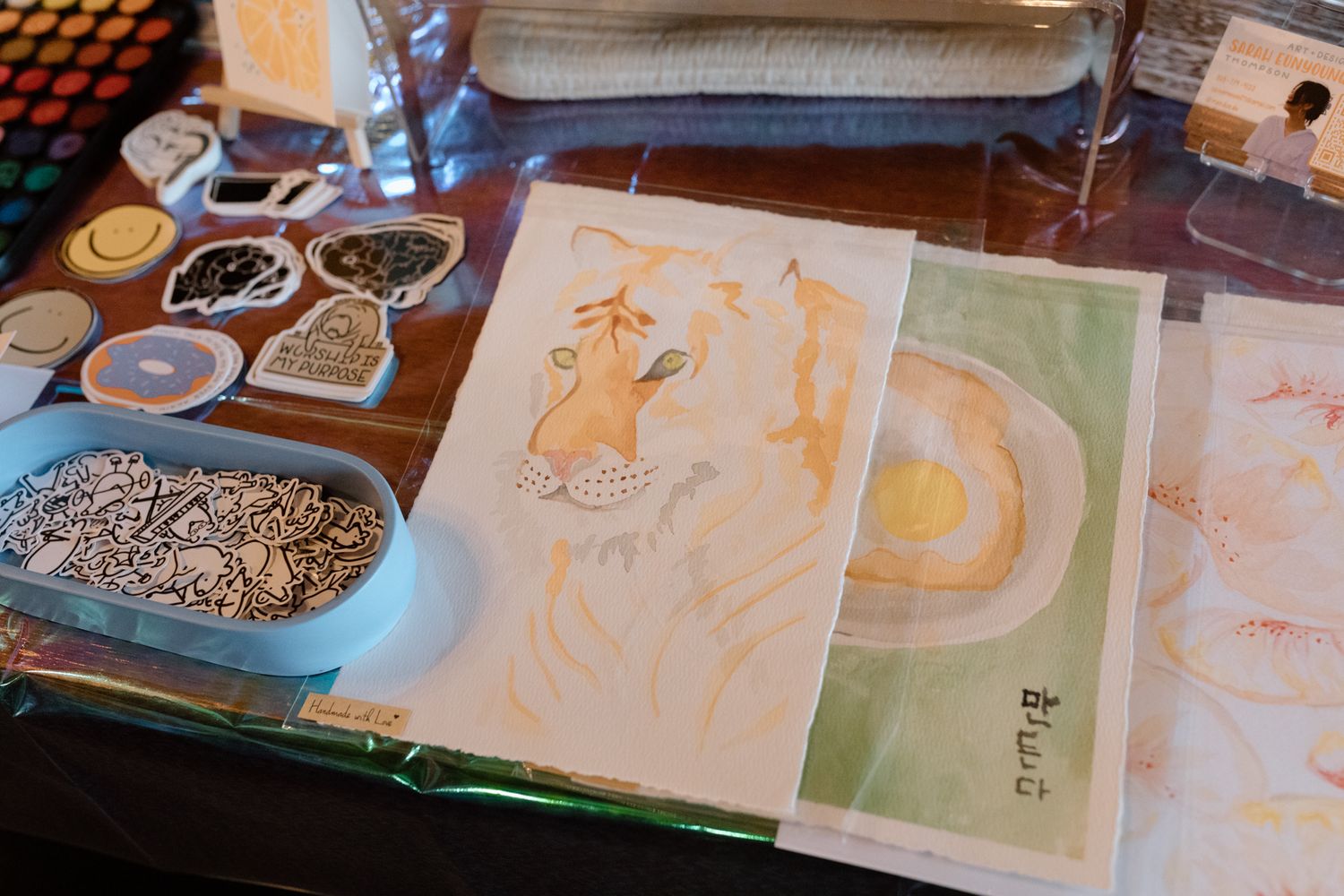 Artist workspace with watercolor paintings, paint palettes, and decorative trays on a glass table.
