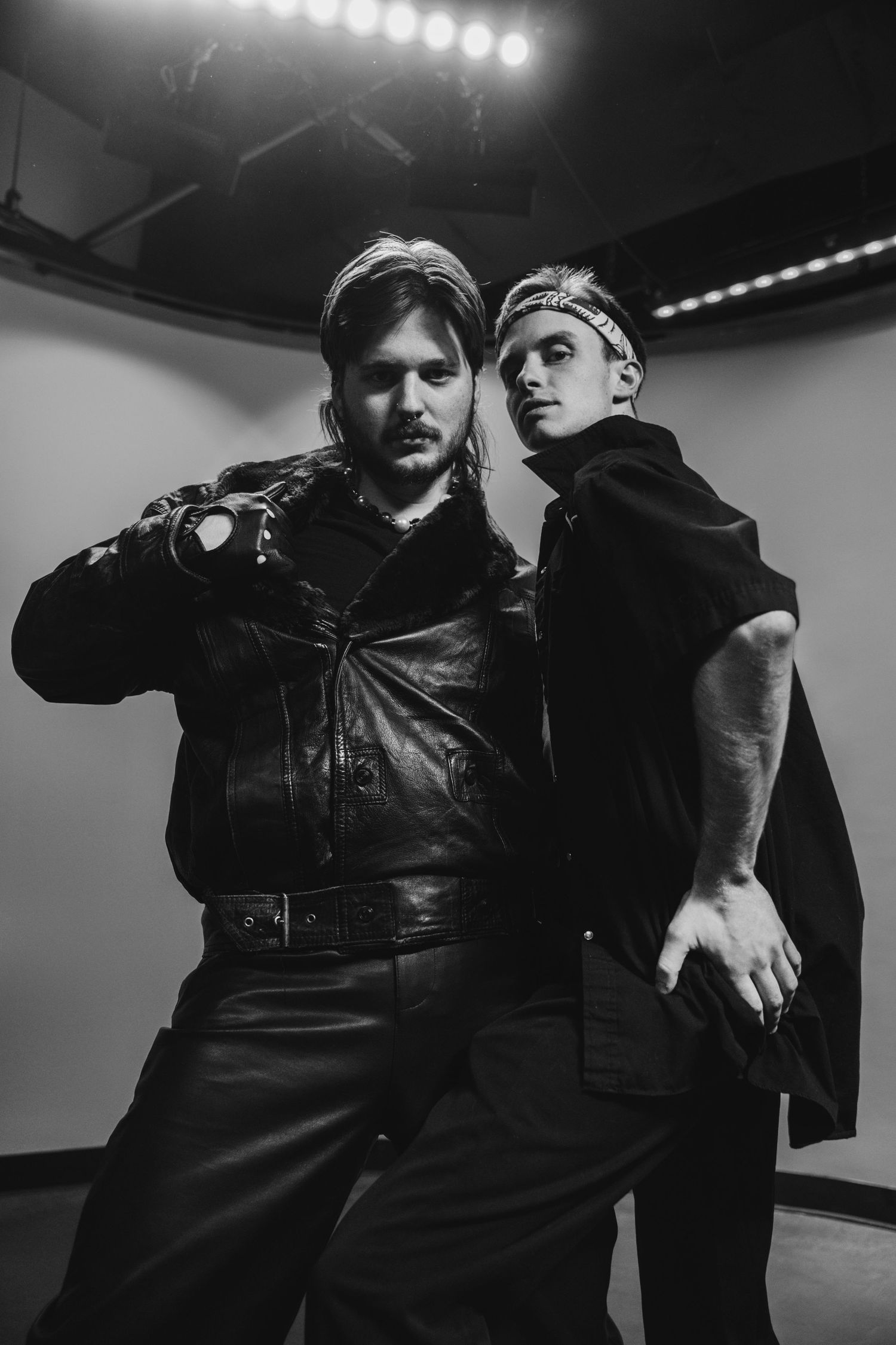 Two men in black leather outfits pose dramatically in a black and white photo shoot under studio lights.
