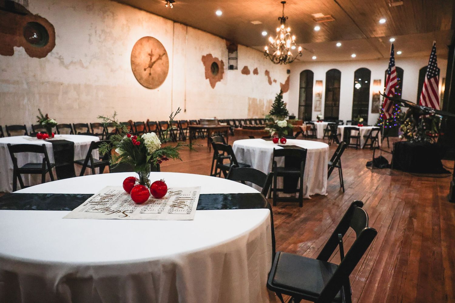Elegant event venue with round tables set with white linens, red roses, and black chairs under warm chandelier lighting.