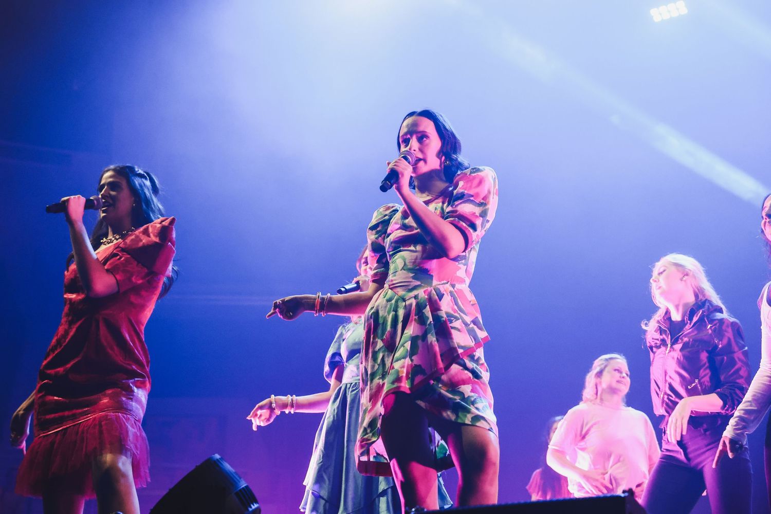 Female singer in floral dress performing on stage with backup dancers under blue concert lights.