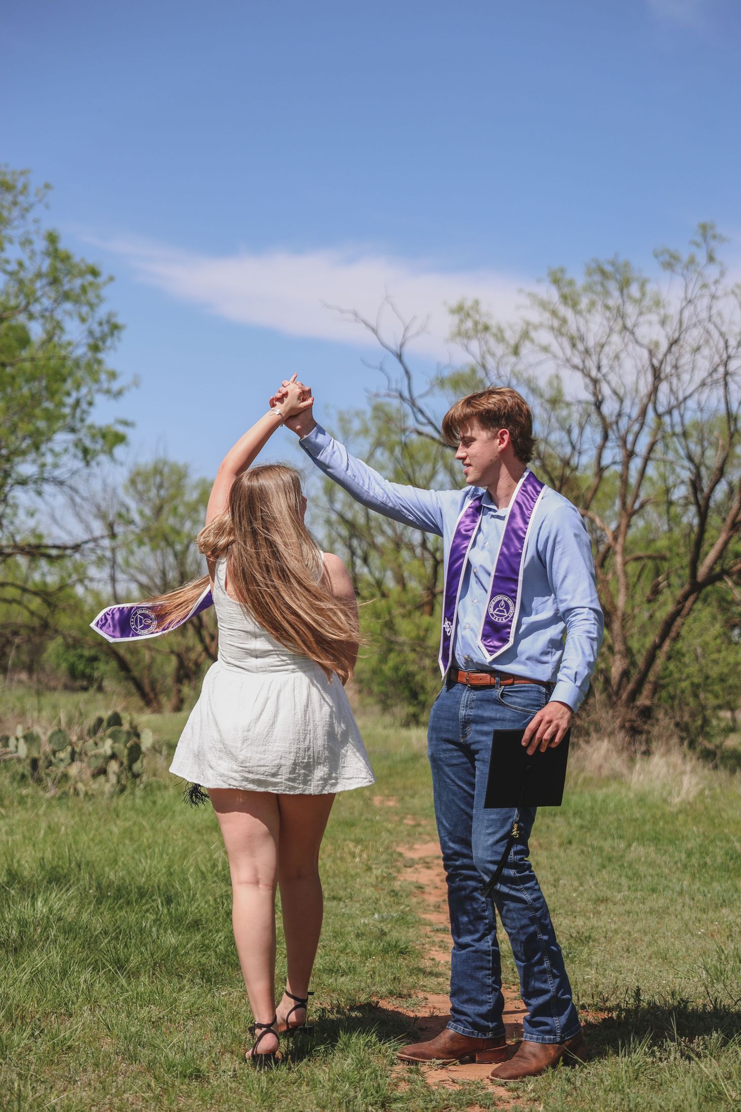 Graduate in purple stole spinning woman in white dress outdoors during spring graduation celebration.