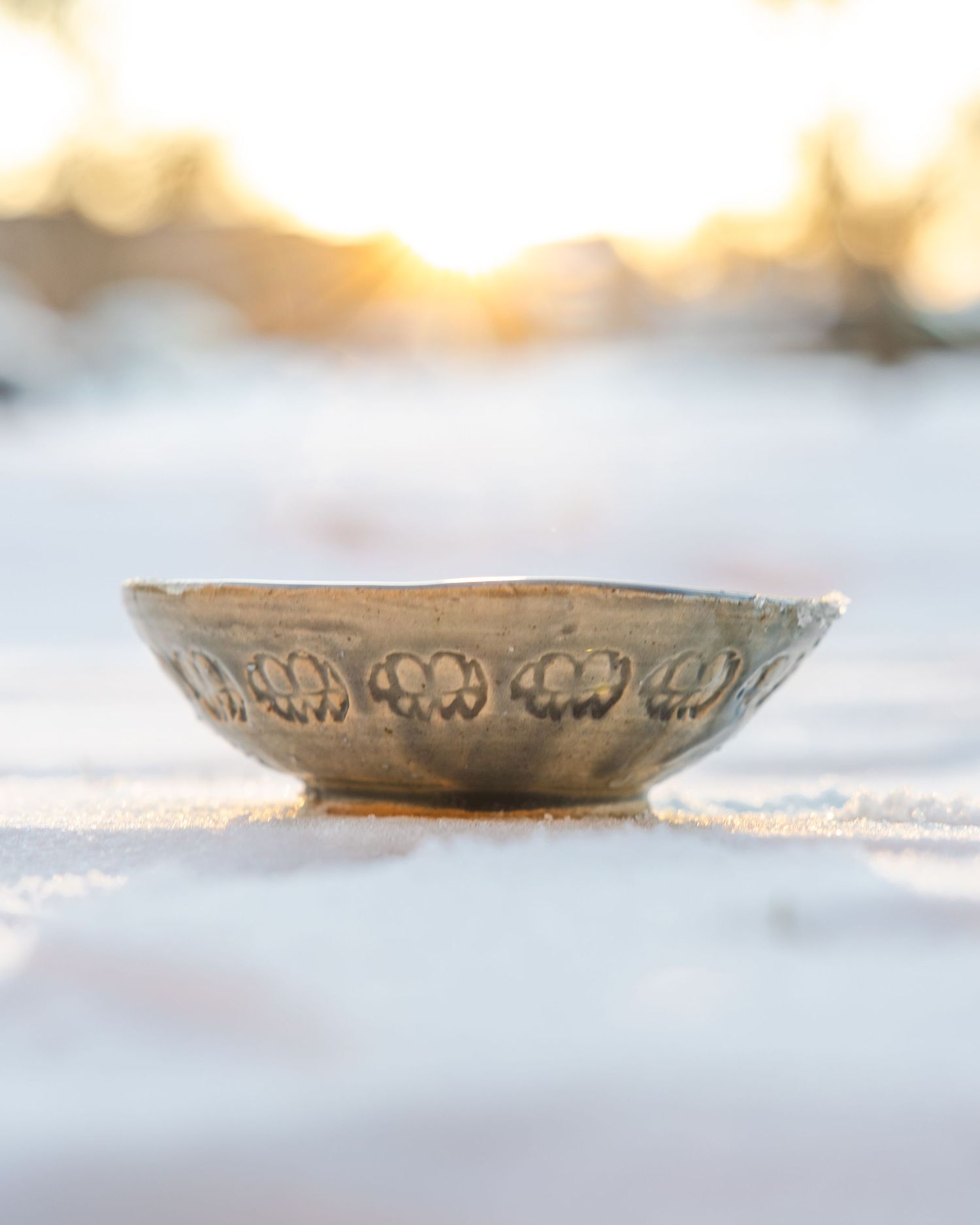 Low shallow ceramic bowl with carved floral design resting on snow at sunset.