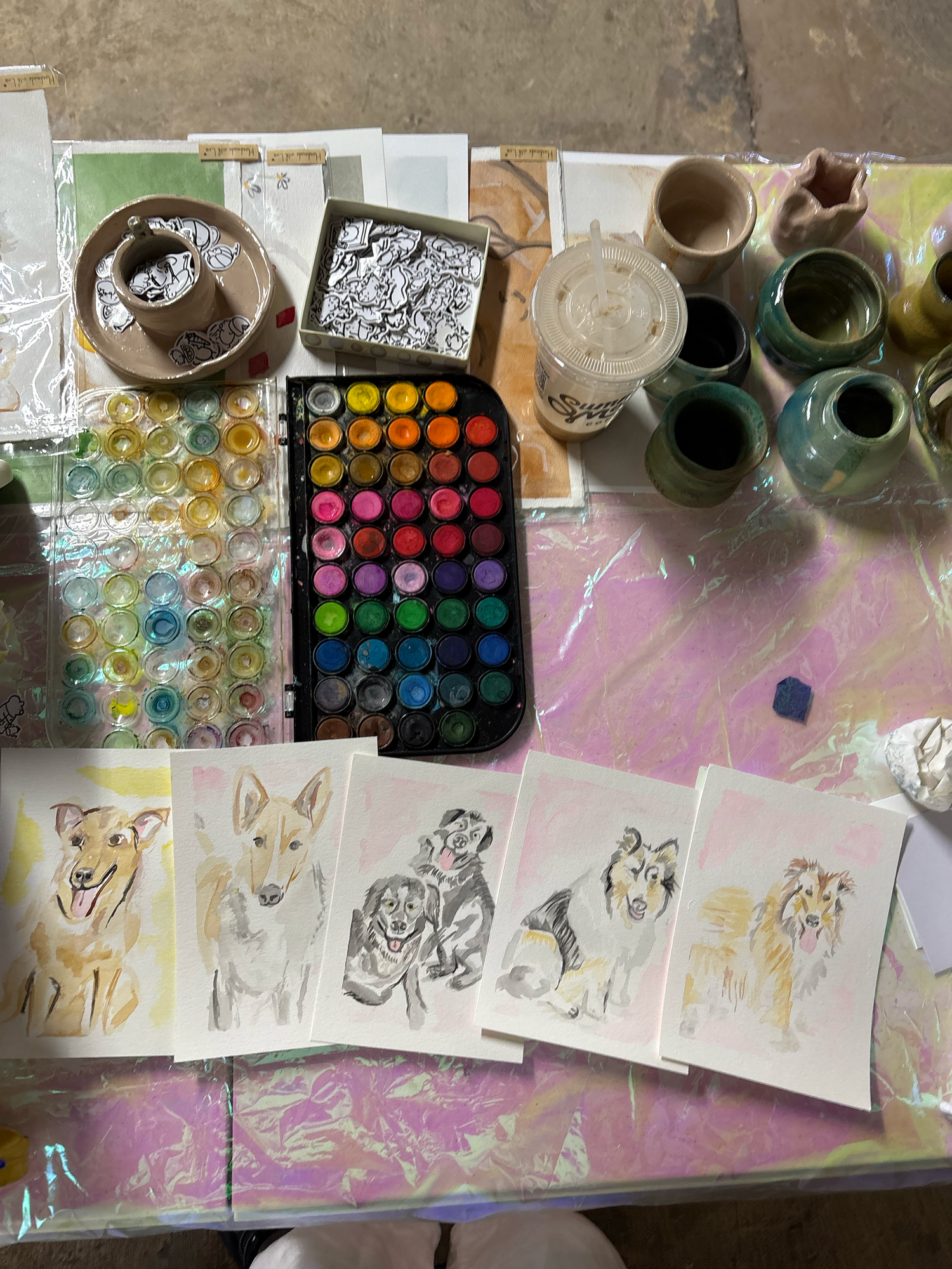 Watercolor painting workspace with dog portraits, colorful paint palette, brushes, and art supplies on a plastic-covered table.