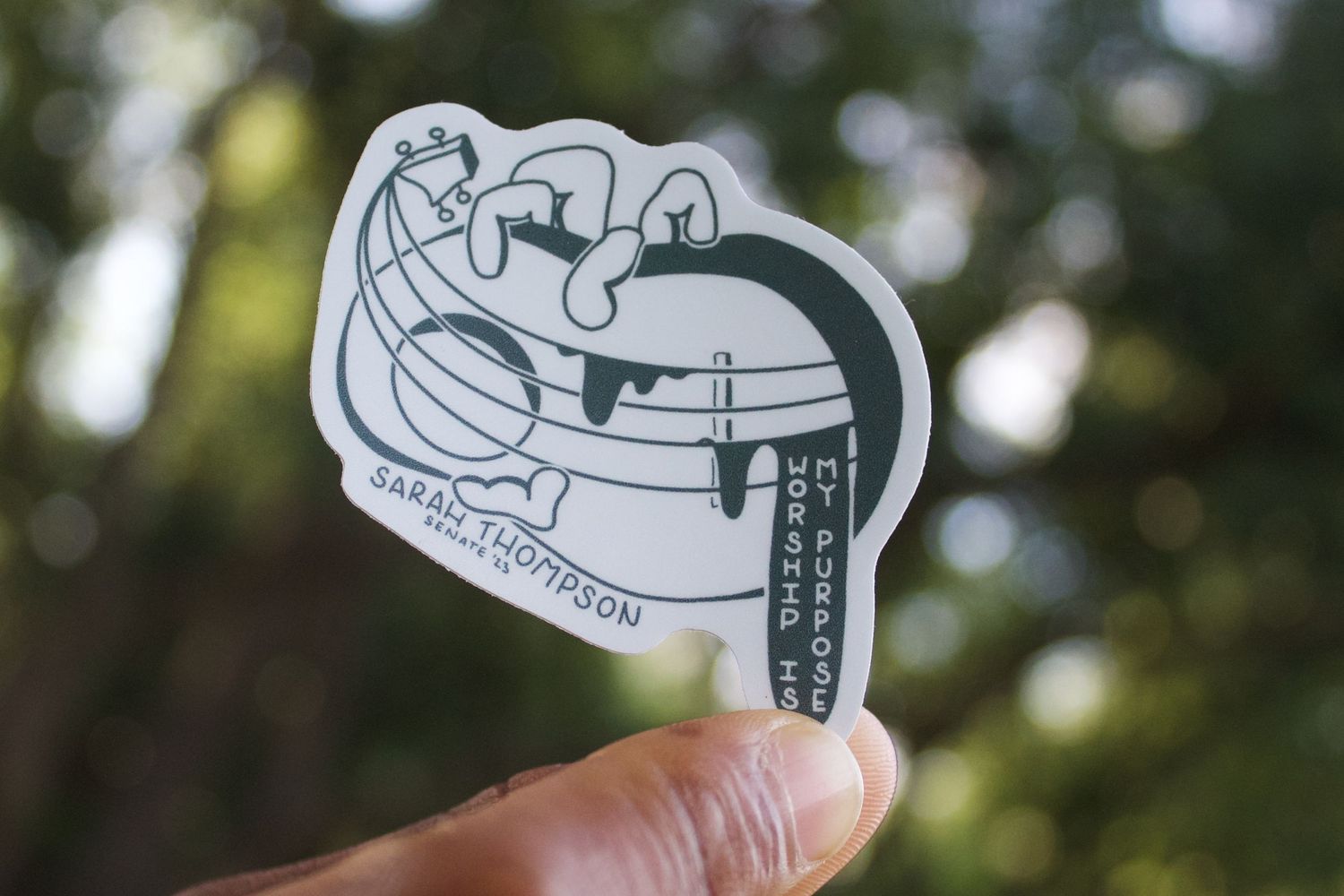 Hand holding a small sticker featuring a globe illustration with Sarah Thompson Monsters Co. branding on a blurred green background.