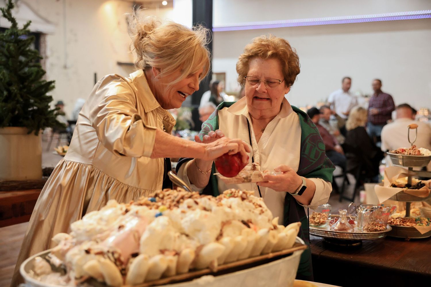 Two elegant older women smiling while serving themselves desserts at a festive indoor catered reception.