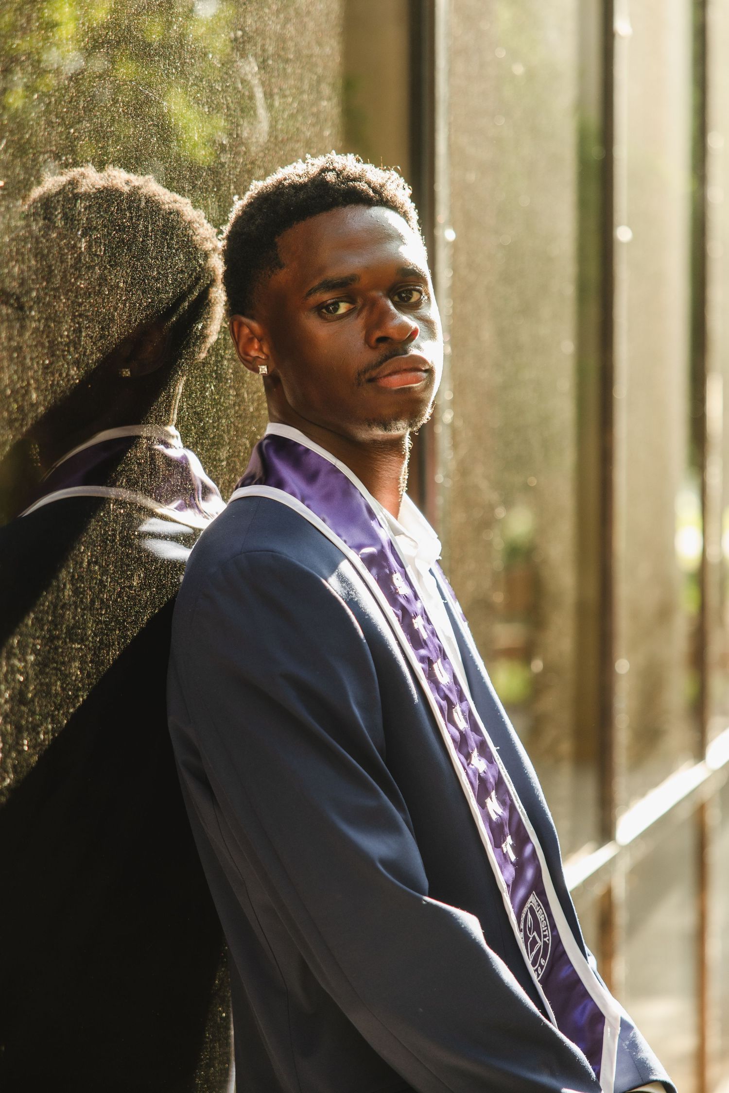 Young Black man in graduation gown with purple stole leaning against reflective wall.