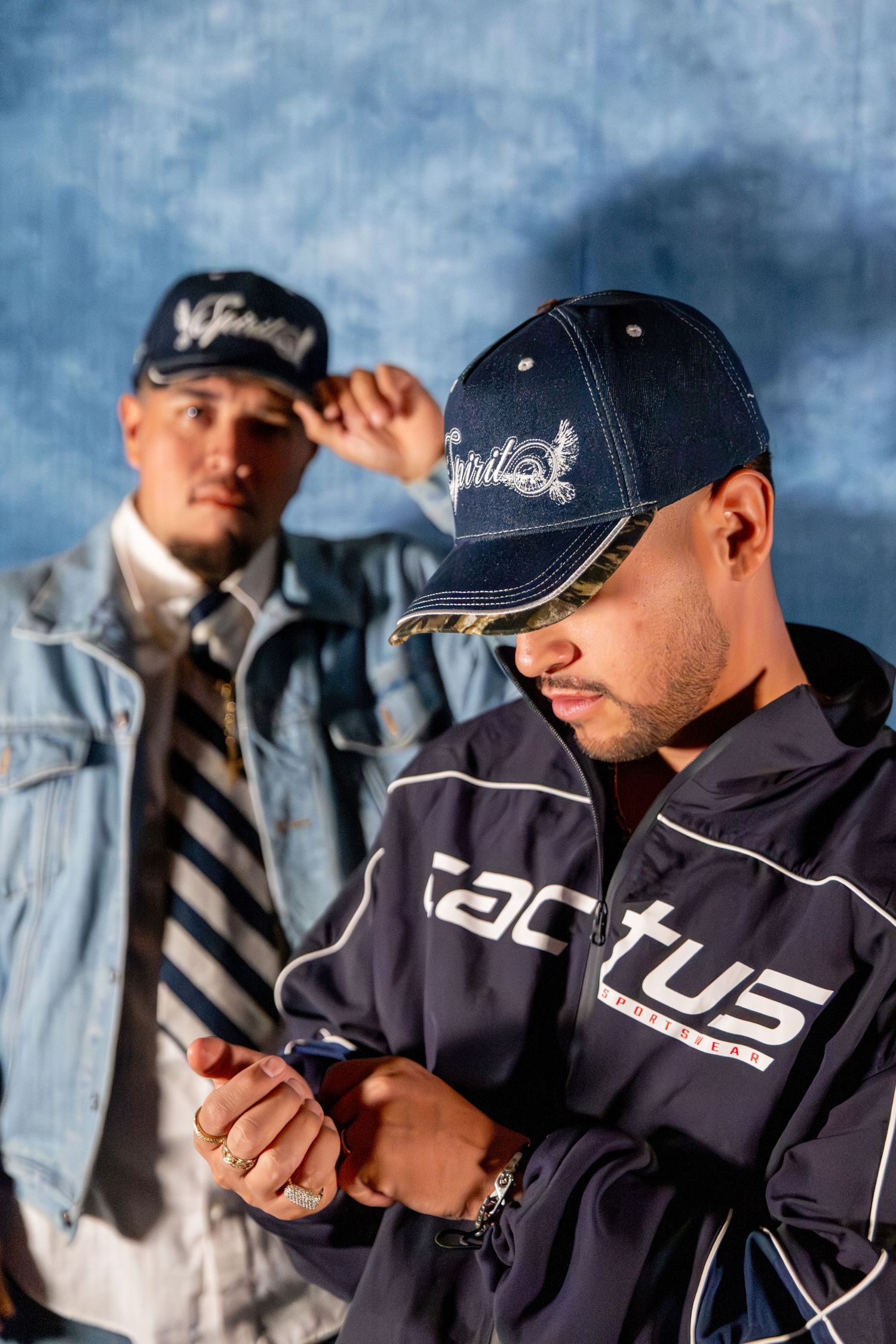 Two men wearing navy caps, one checking a phone, pose against a blue background.