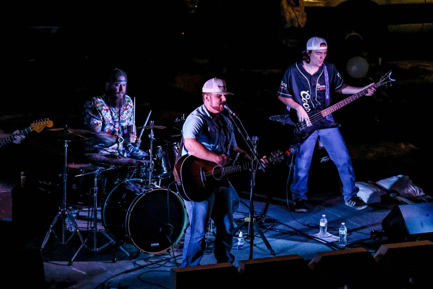Three-piece band performing live on stage with guitarist, drummer, and bassist under blue lights.