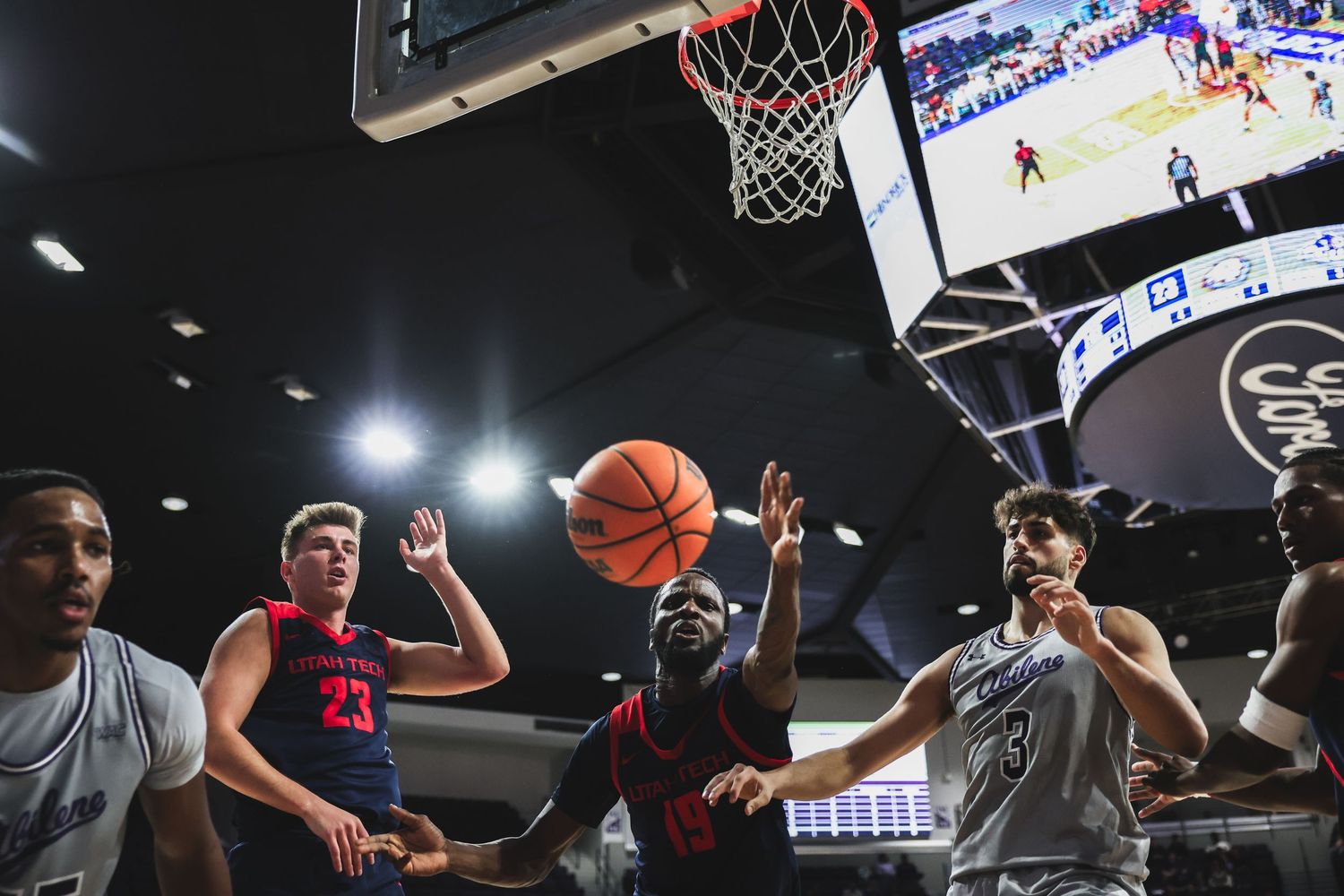 College basketball players from two teams compete for a rebound under the basket in an indoor arena.