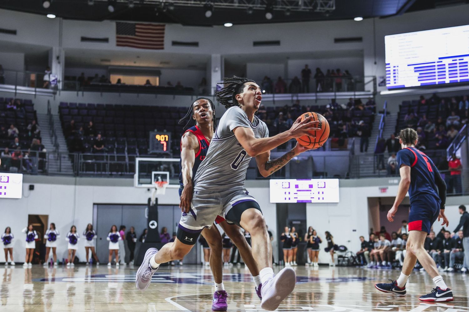 College basketball player in gray uniform drives to the basket during an indoor arena game.