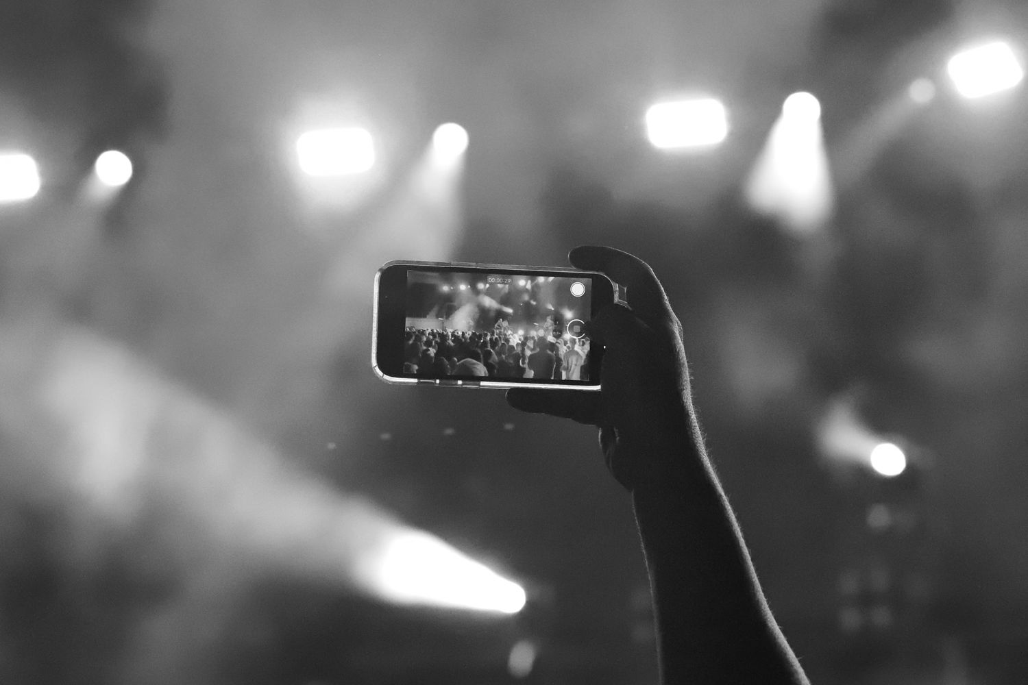Black and white photo of a hand holding a smartphone recording a live concert with stage lights.