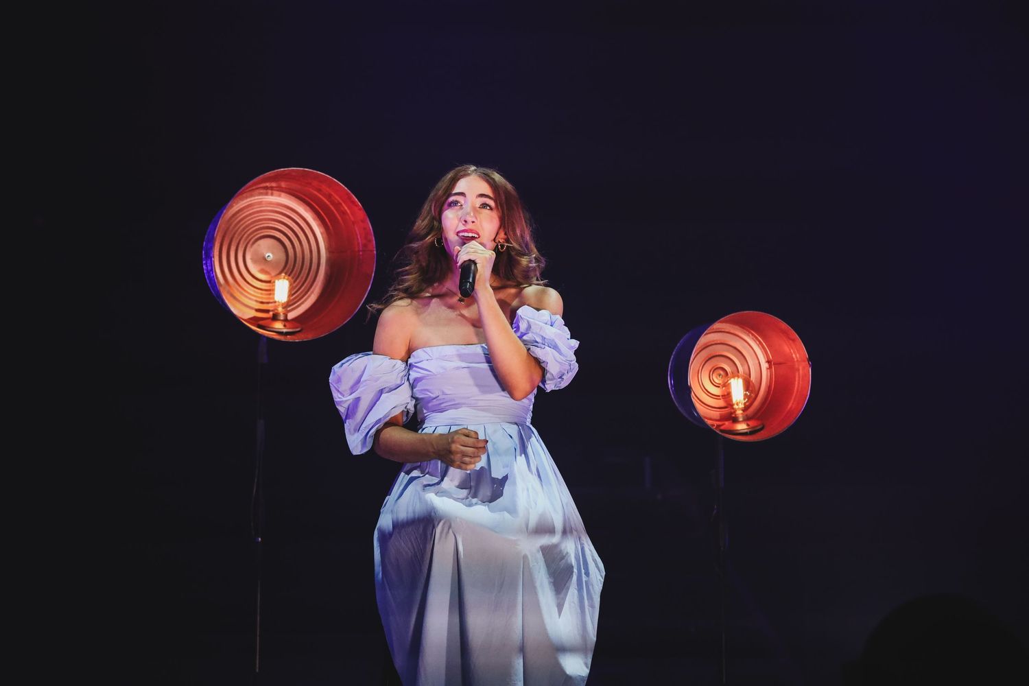 Female singer in white off-shoulder dress performing solo on stage with red circular lights.