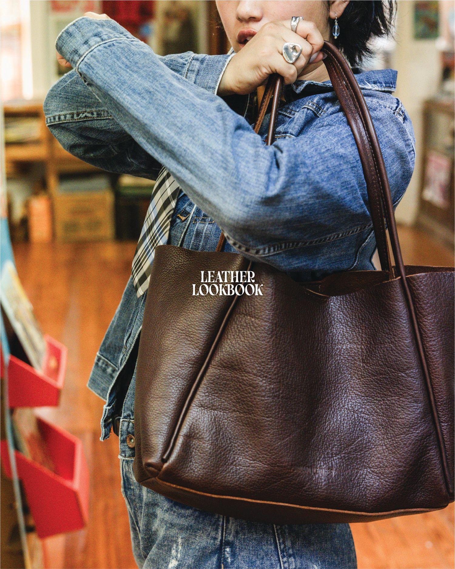 Person wearing denim jacket holding a large dark brown leather tote bag, featured in Leather Lookbook.