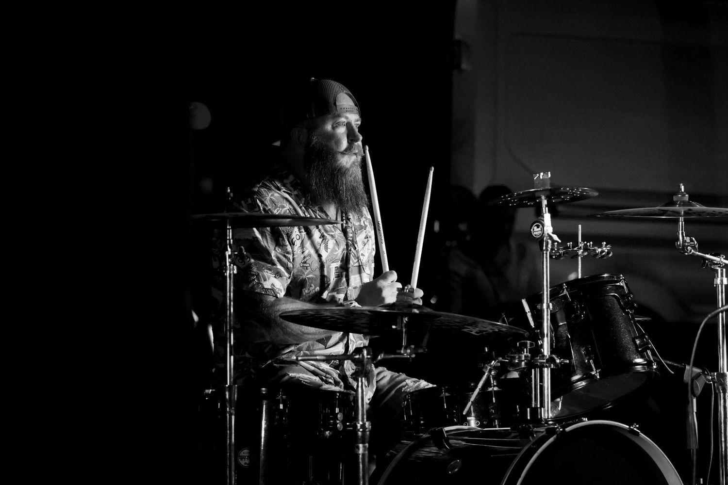 Black and white close-up of a long-haired drummer playing drums intensely at a live music performance.