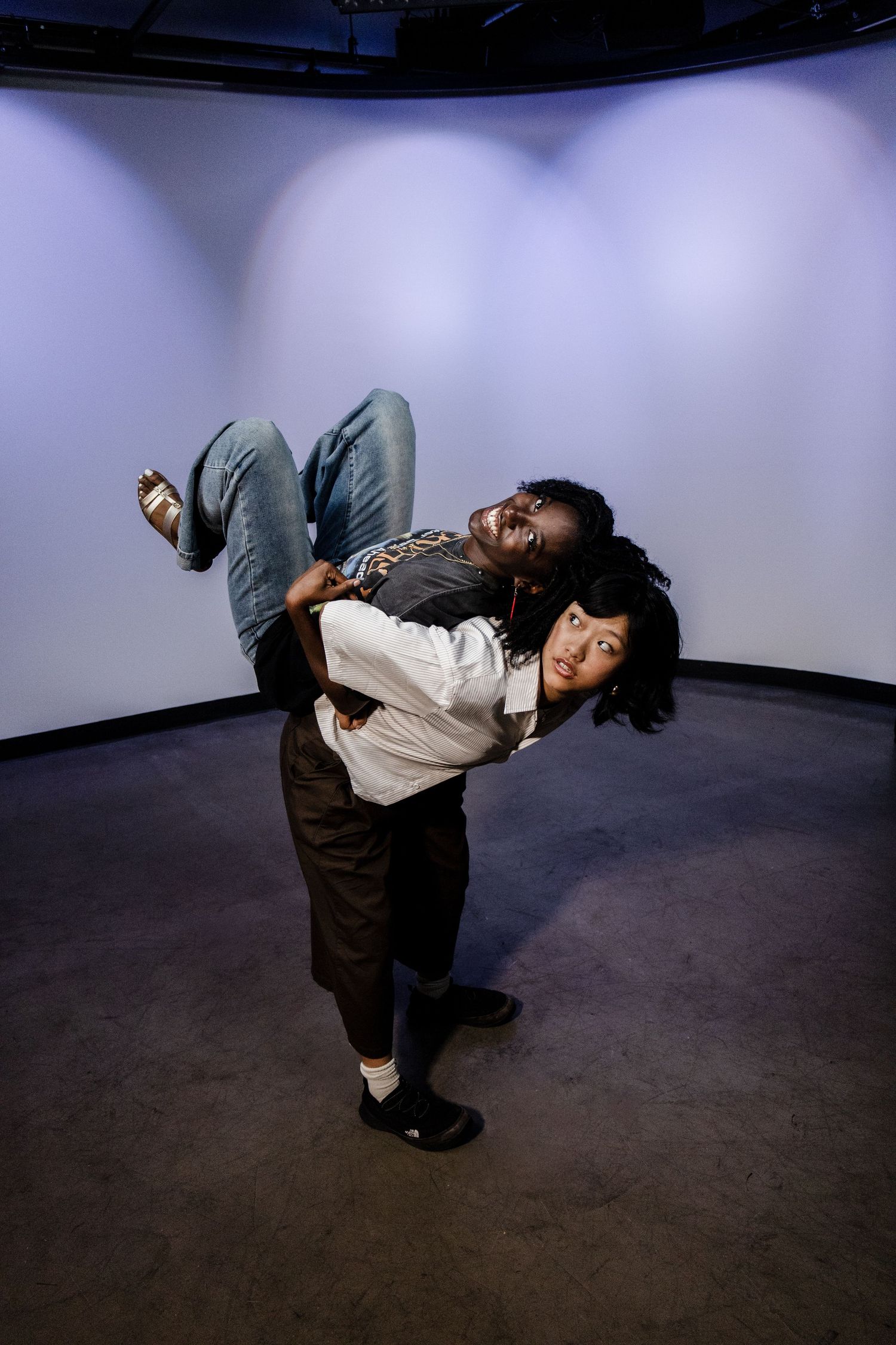 Woman carries man on her back in playful pose inside a curved white studio space.