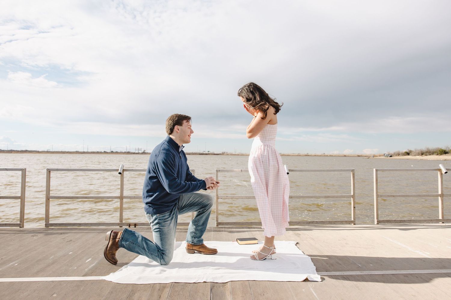 Man proposing on one knee to woman on a waterfront dock under a cloudy sky.
