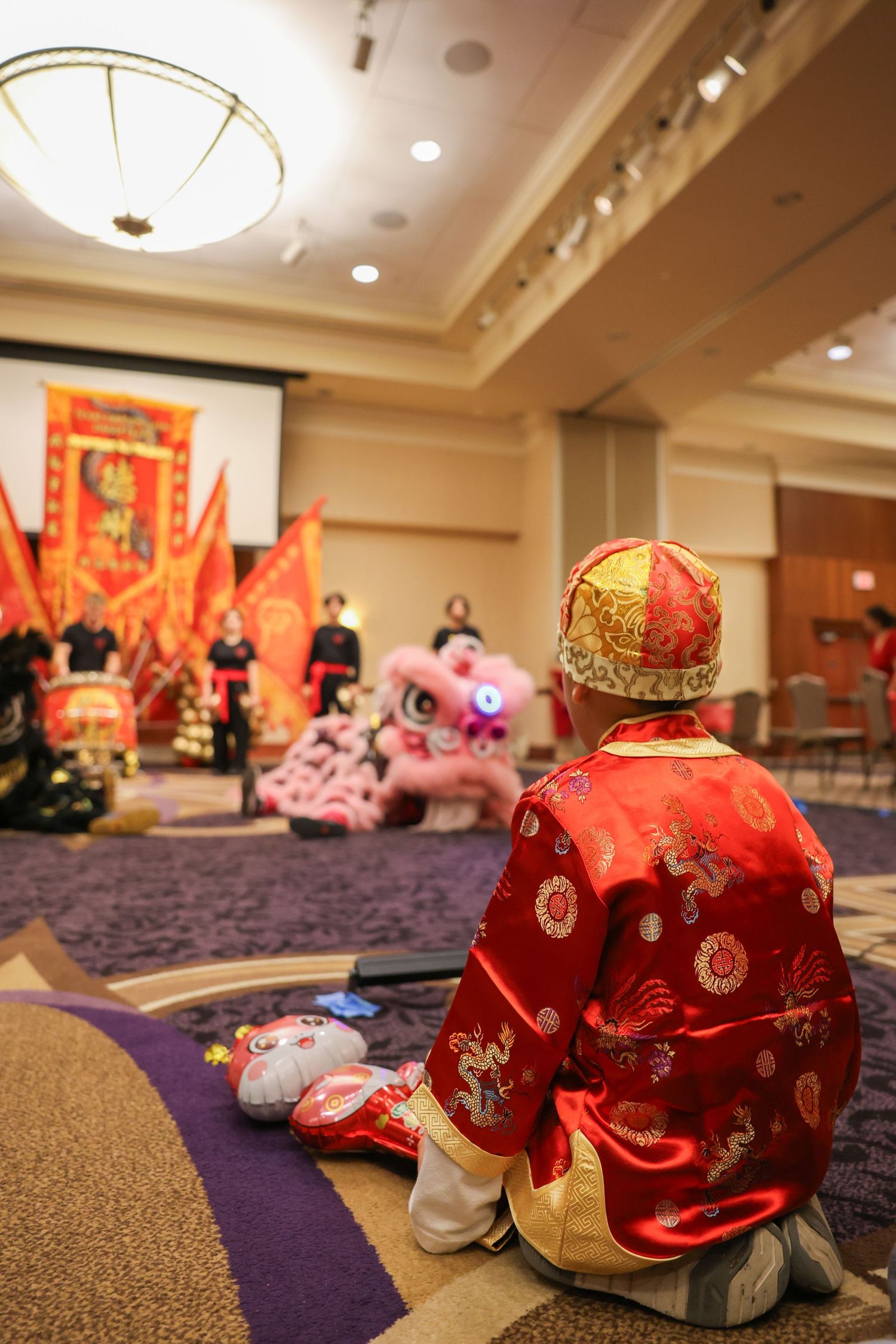 Child in traditional red Chinese robe watches colorful lion dance performance in hotel ballroom.