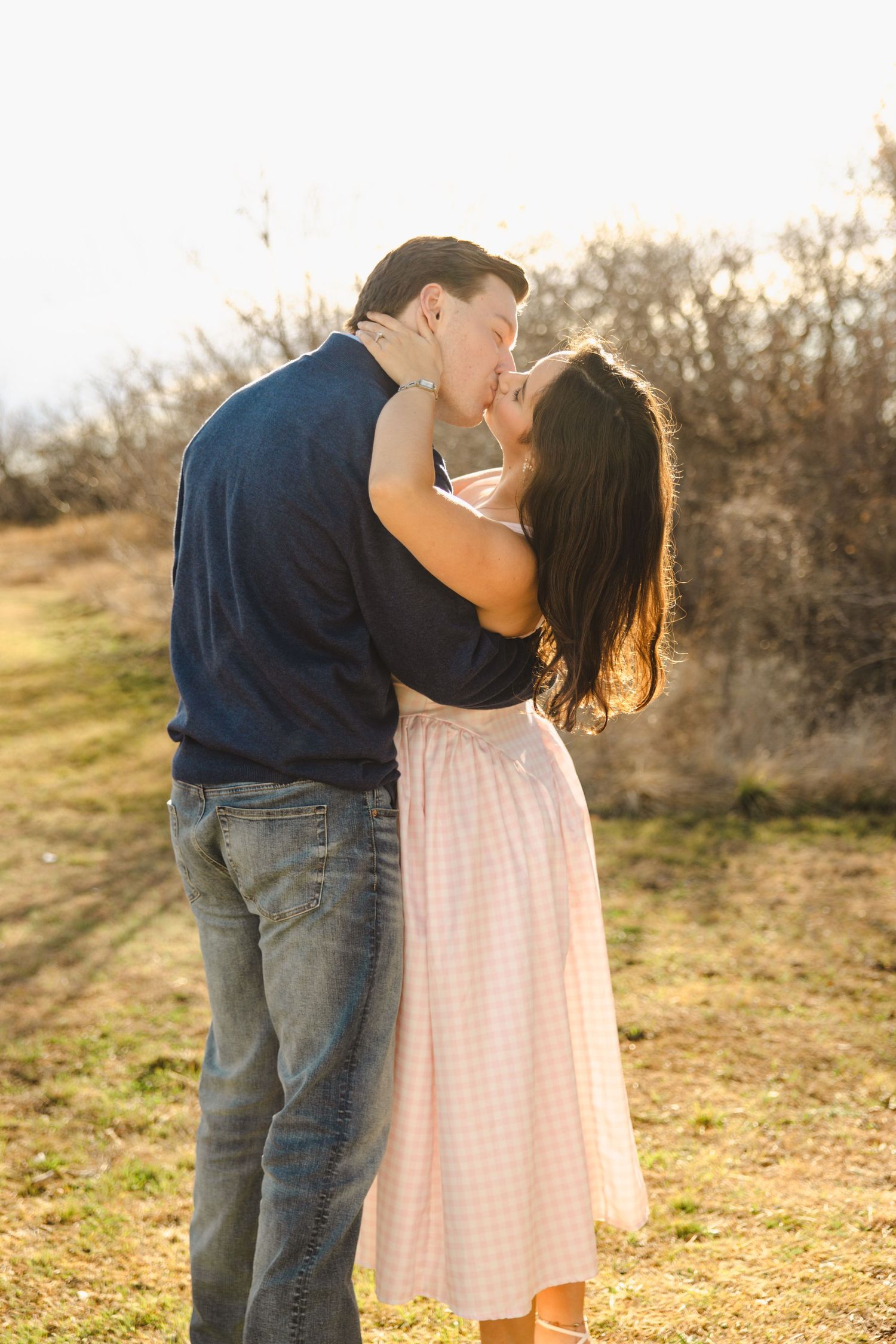 Couple sharing a romantic kiss outdoors in a sunny field with dry brush background.