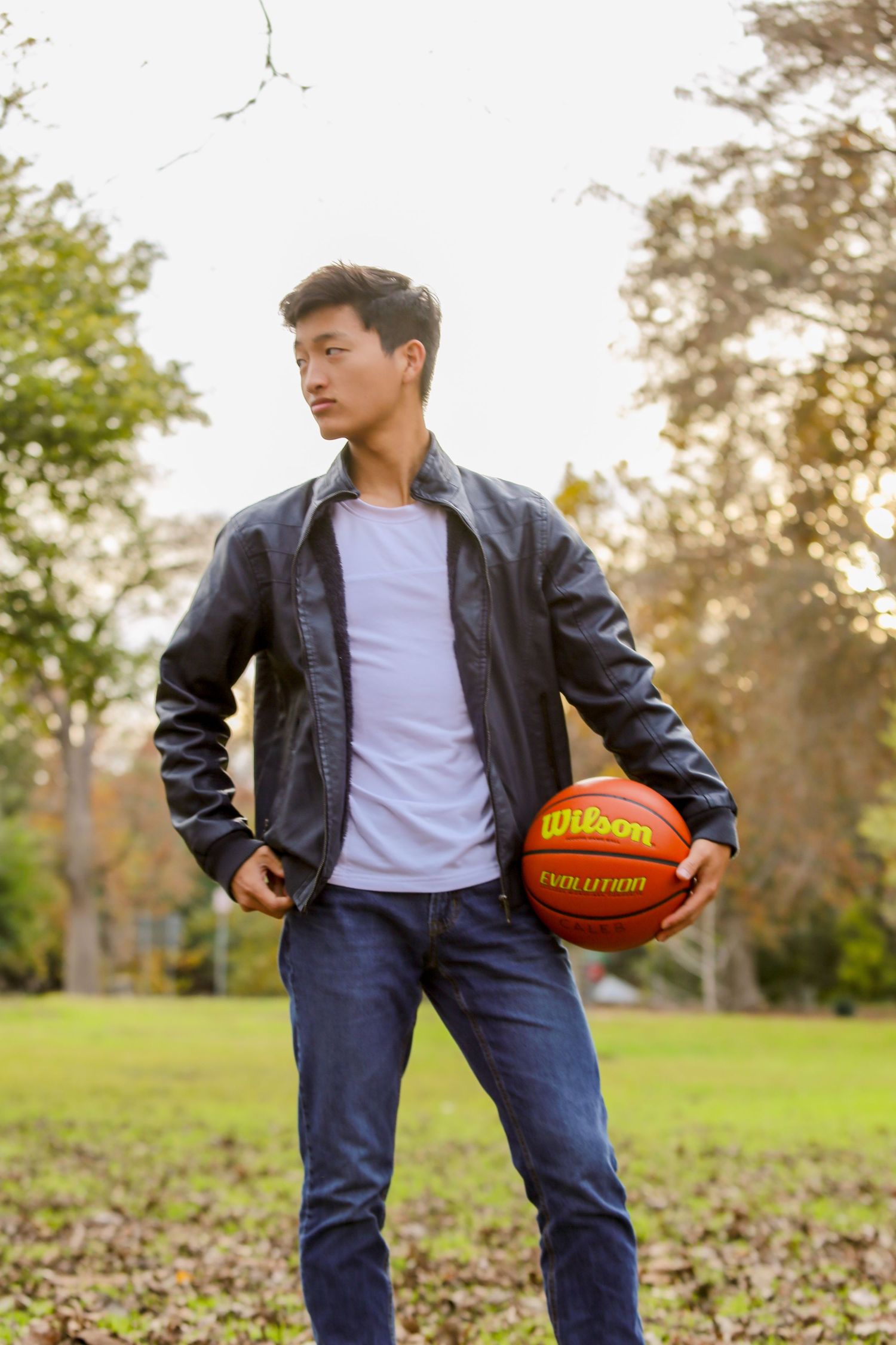 Young man in black leather jacket holding a Wilson basketball in an autumn park setting.