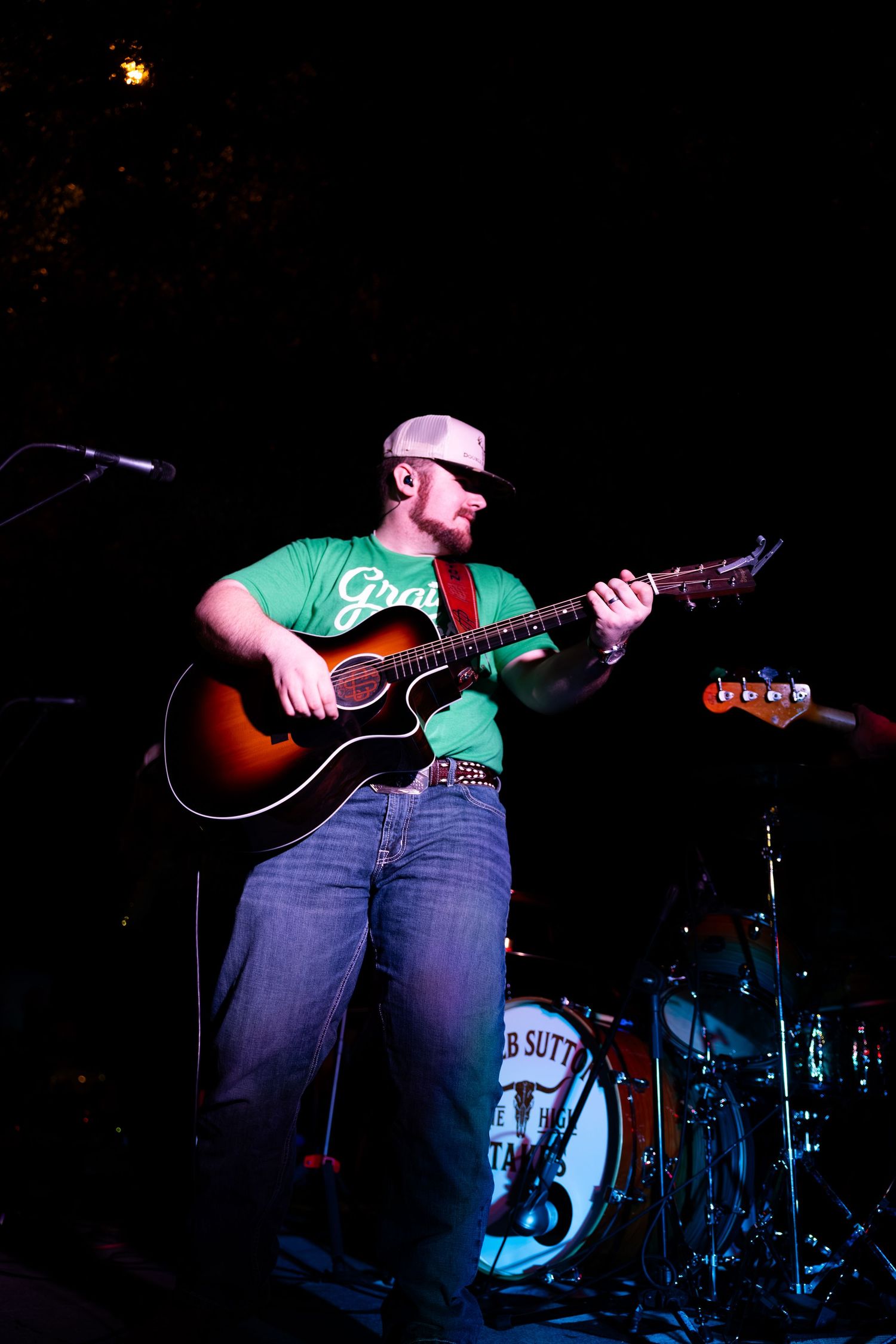 Man in green t-shirt and cap playing acoustic guitar on dark stage during live performance.