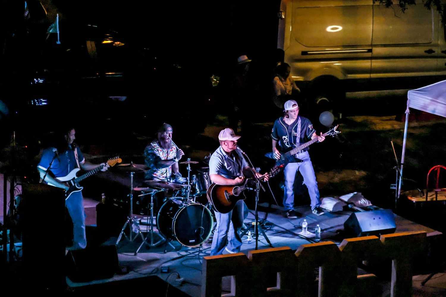 Four-piece band performing on an outdoor stage at night with a van visible in the background.