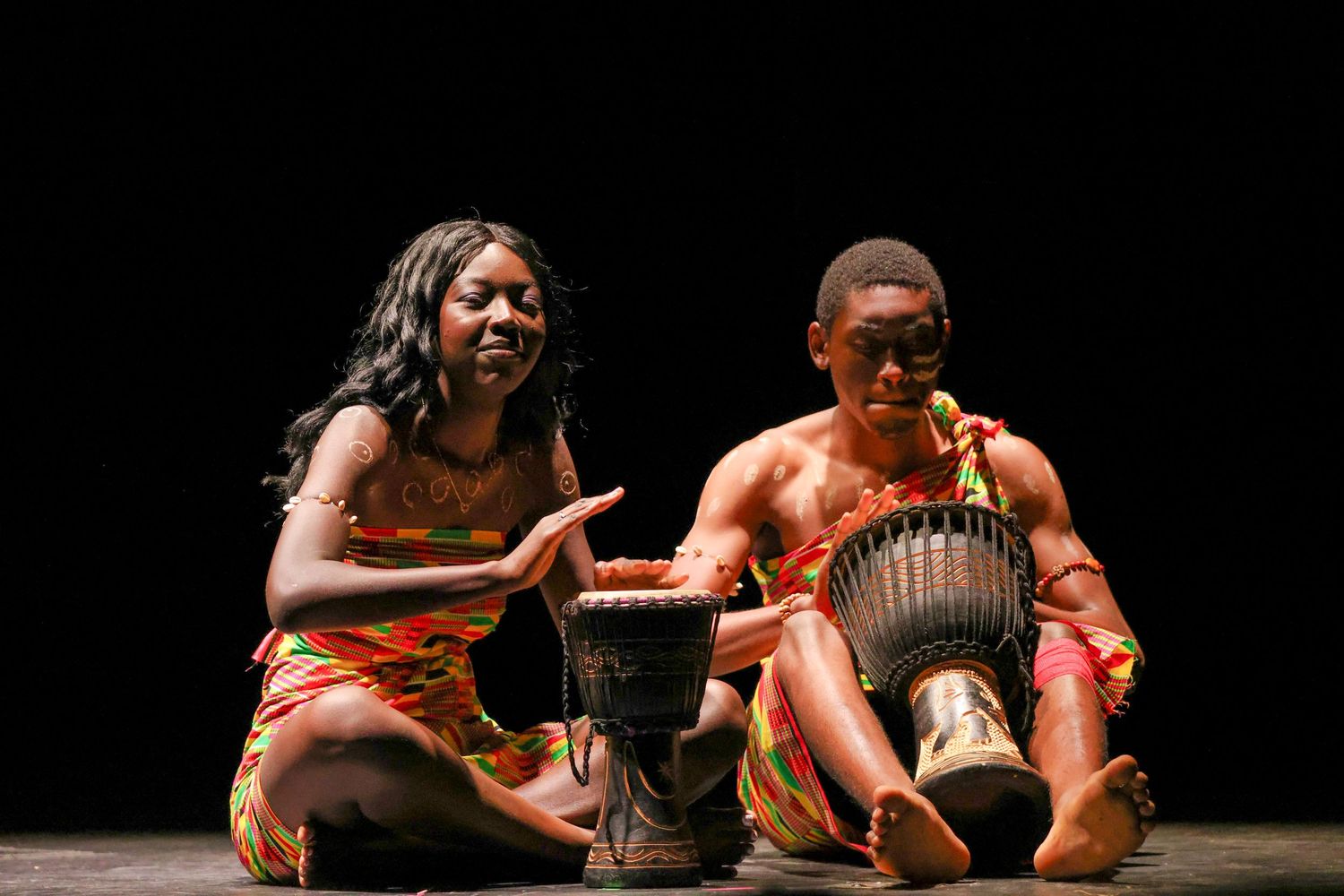 Two African performers in traditional attire play djembe drums during a vibrant cultural stage performance.