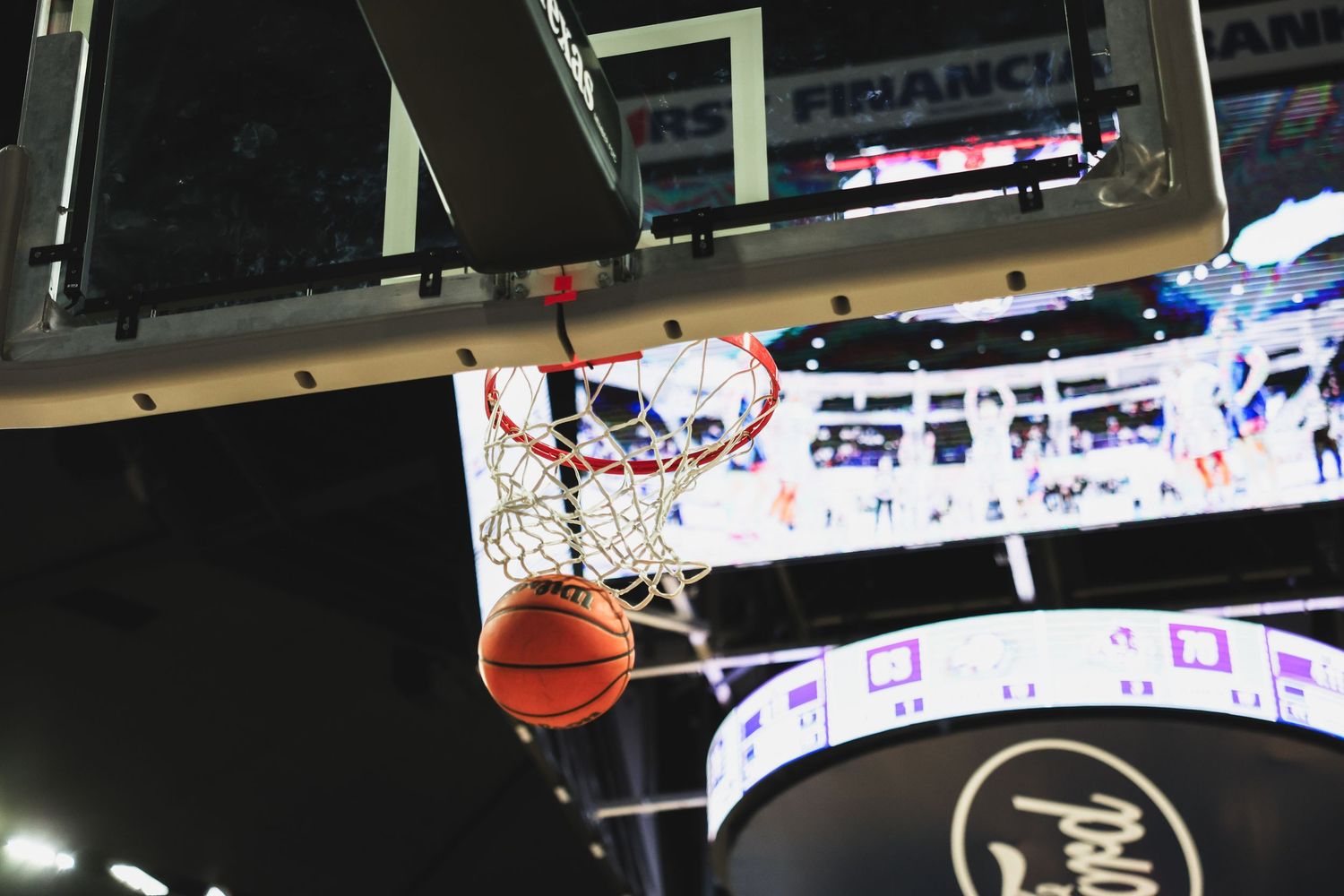 A basketball passes through the net from below in a dramatic close-up shot inside a college basketball arena.