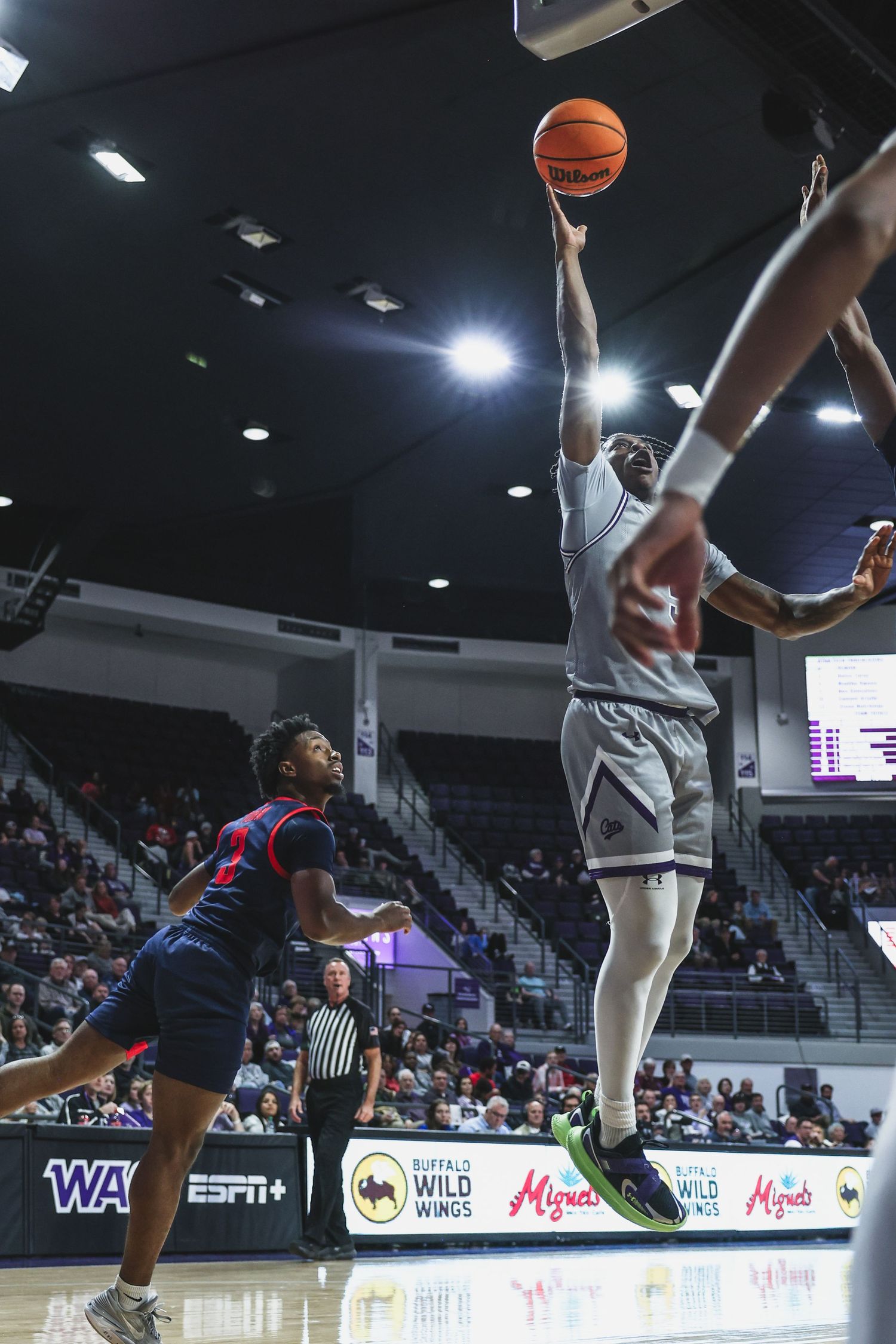 Basketball player in gray uniform shoots a layup over a red-uniformed defender in a college game.