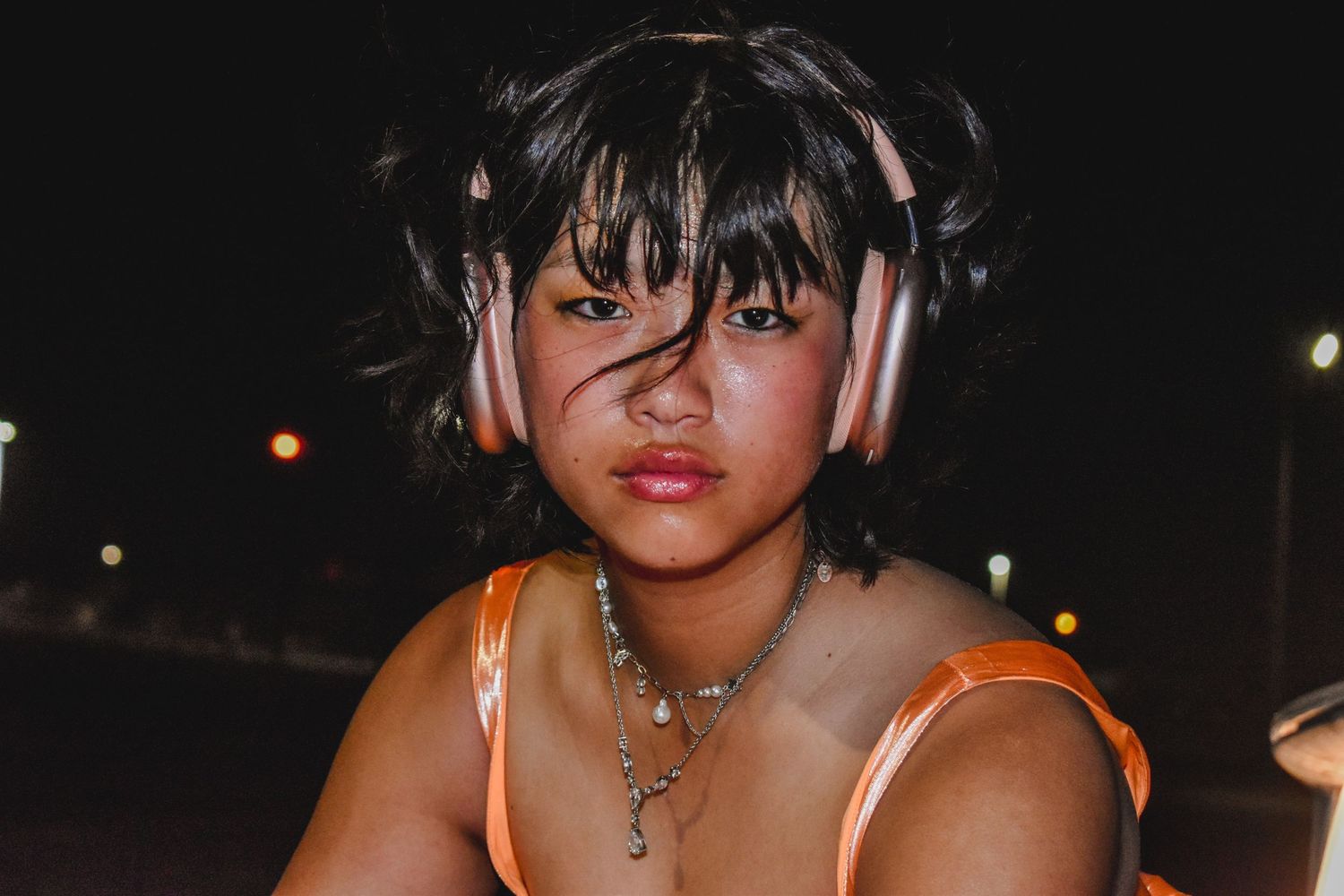 Young woman wearing pink headphones and orange top poses outdoors at night.