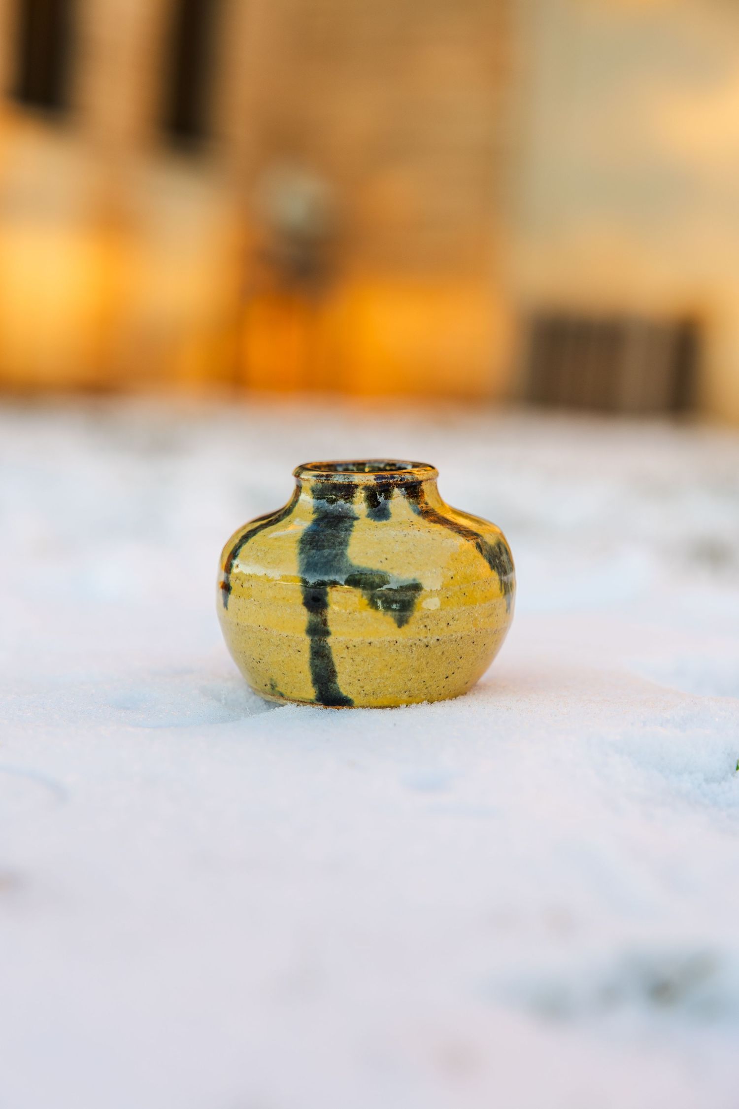 Small yellow ceramic vase with dark brushstroke glaze design sitting on snow.