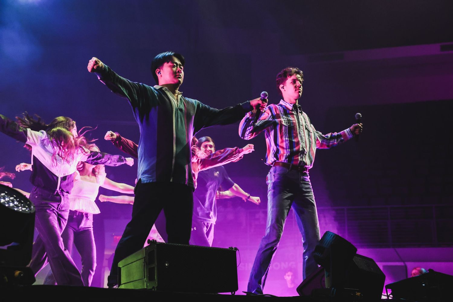 Two male performers singing on stage with dancers behind them under purple concert lighting.