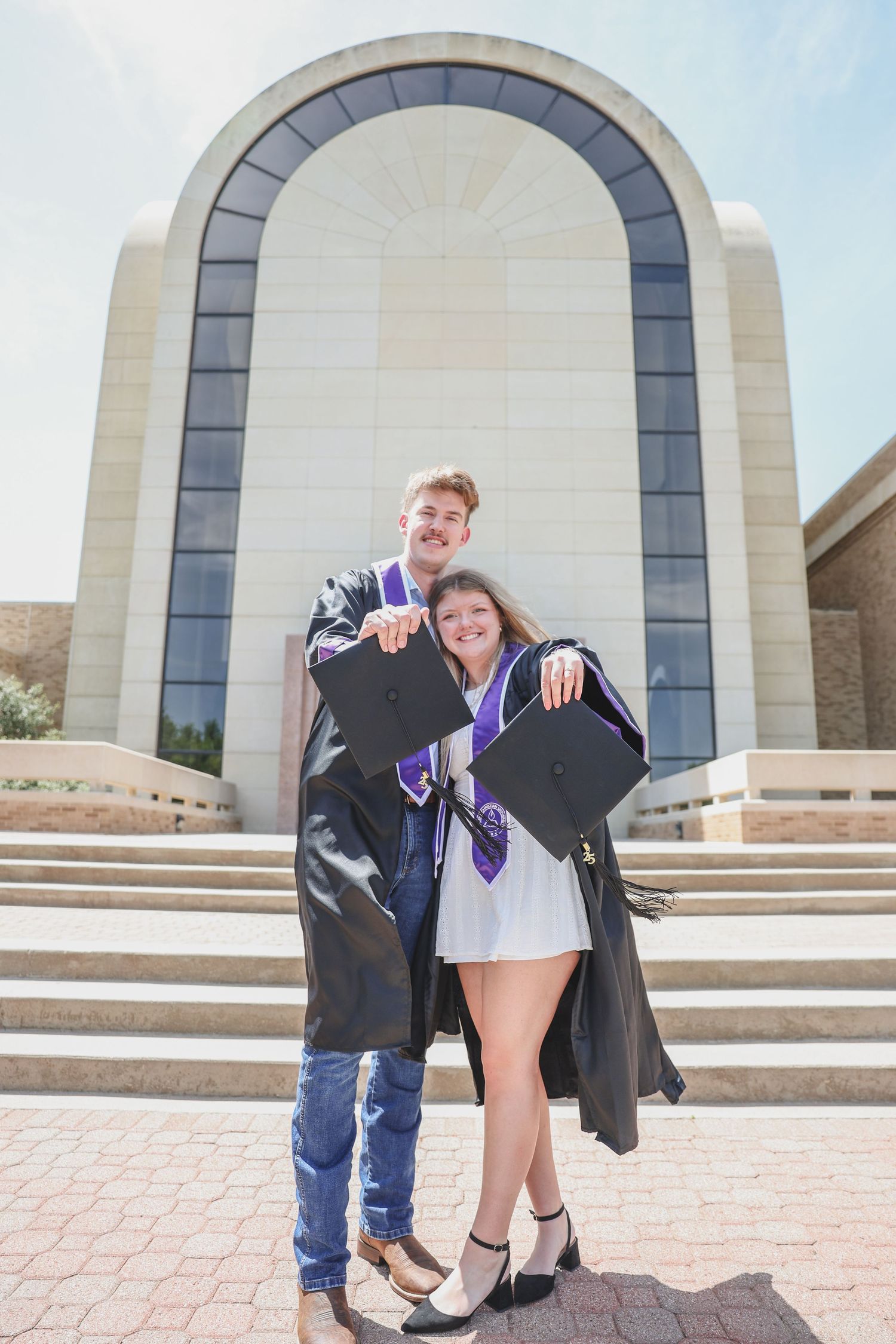 Two graduates in caps and gowns with purple stoles posing proudly in front of a university building.