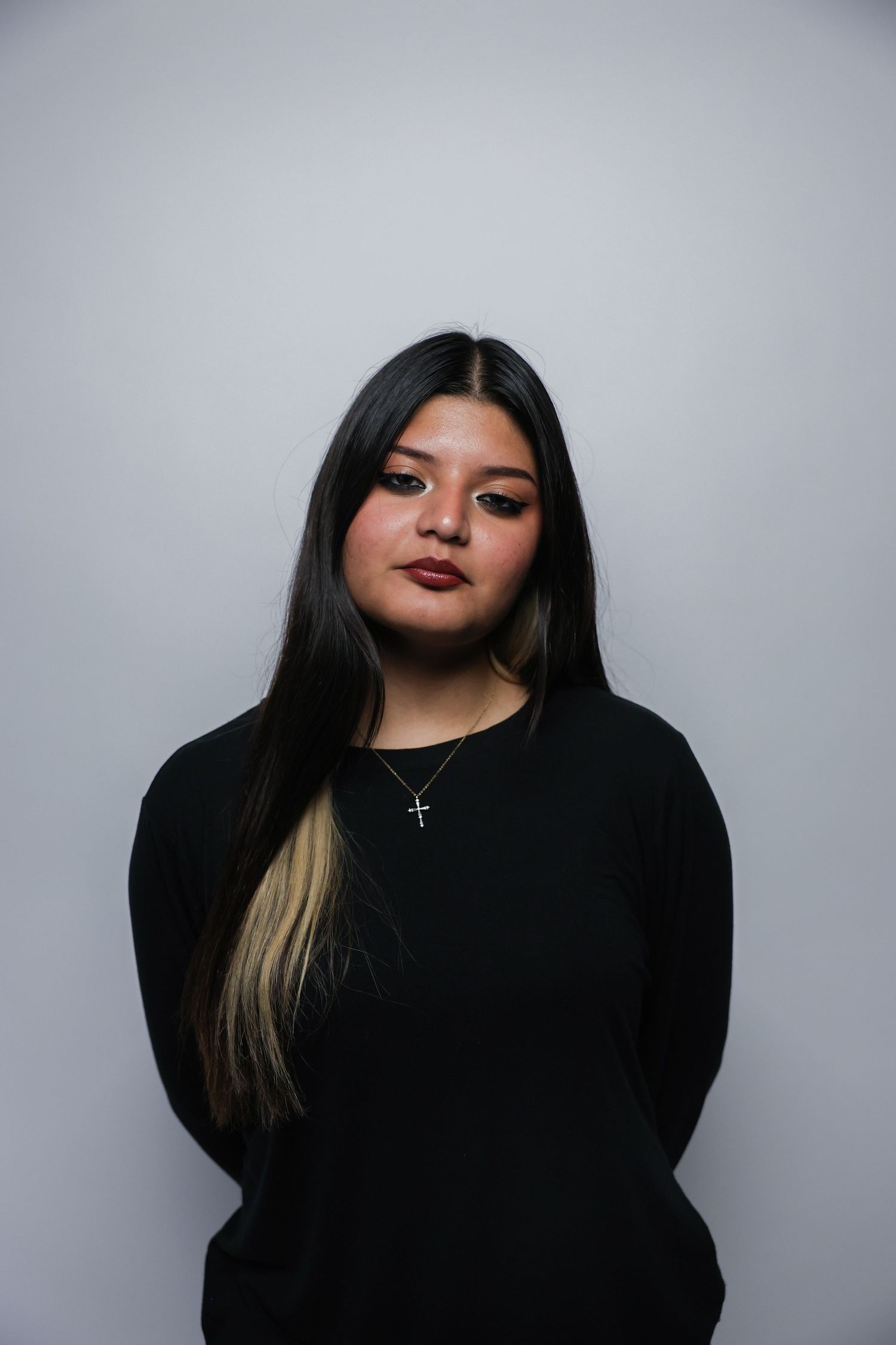 Young woman with long dark hair wearing a black sweater and cross necklace poses against a gray background.