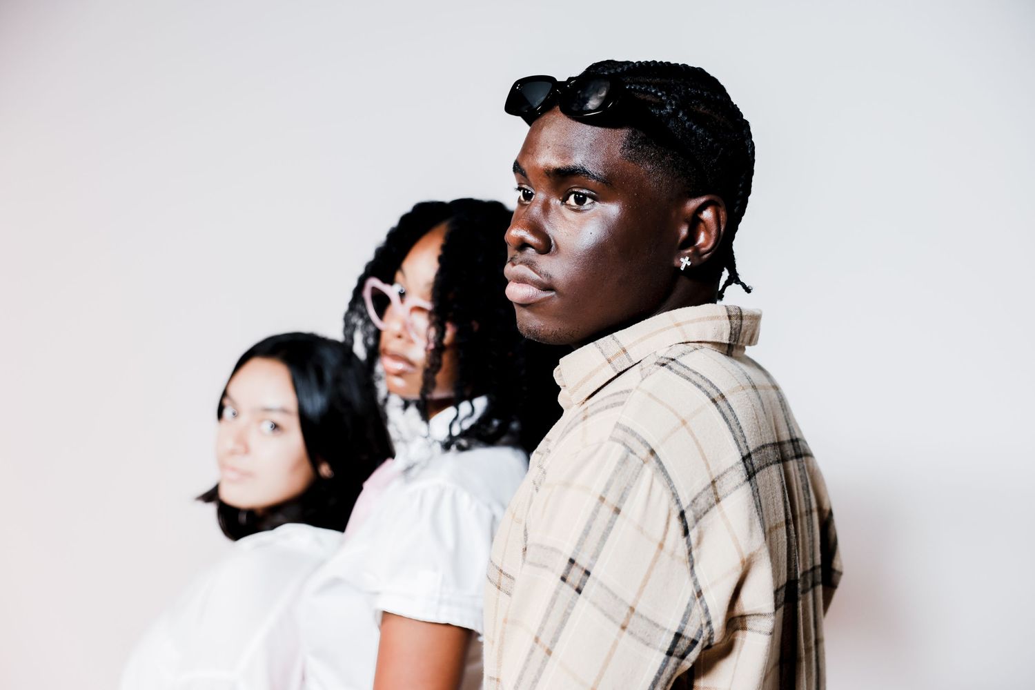 Three diverse young people posing together, man in plaid shirt in foreground against white wall.