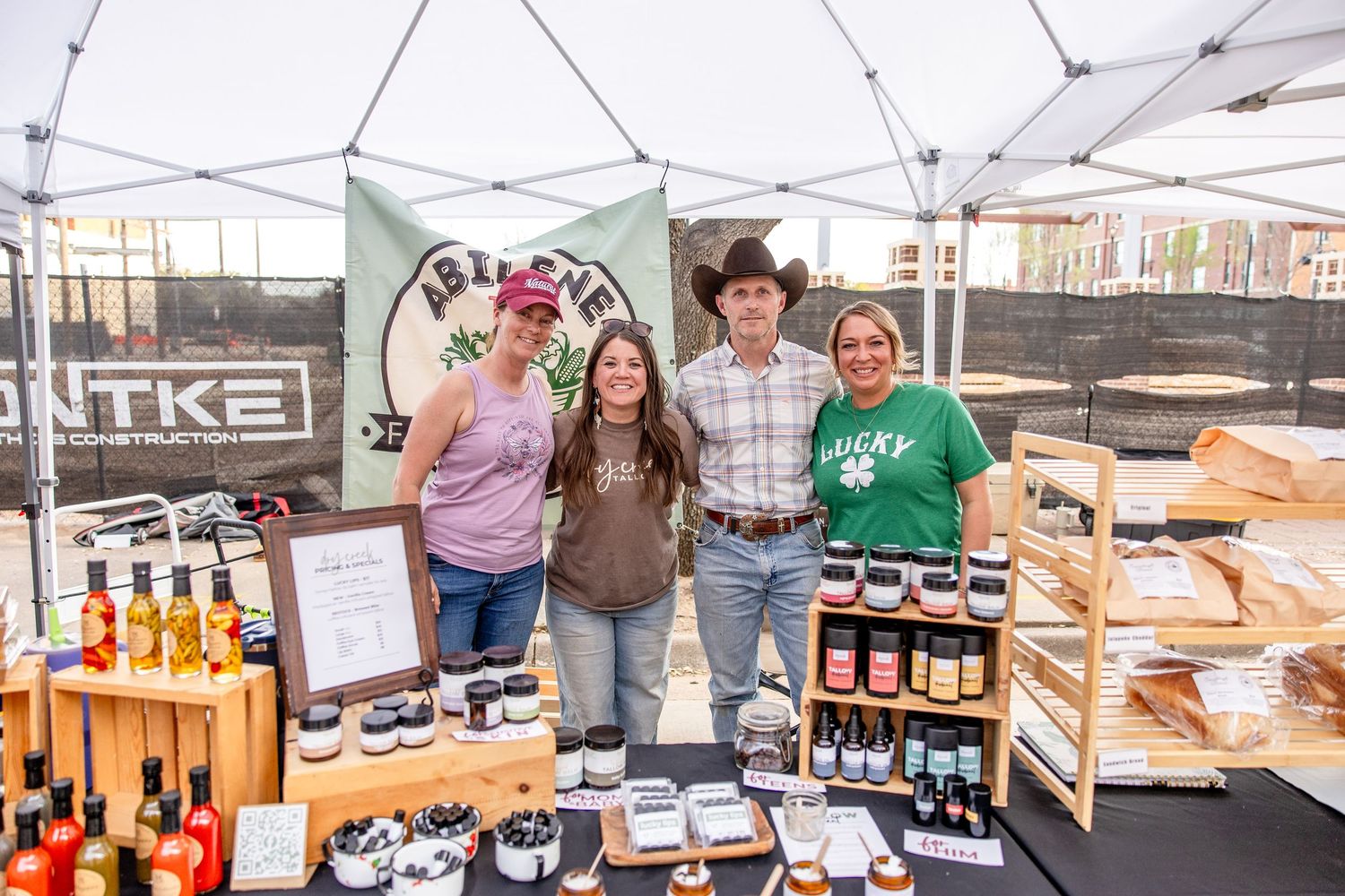 Four people smiling at a vendor booth displaying hot sauces and jams at an outdoor market.
