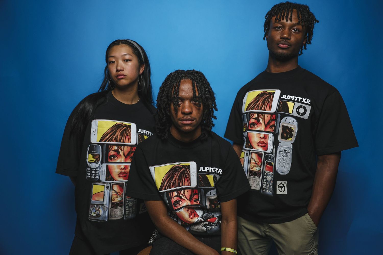 Three young people wearing matching black JUPITTXR graphic tees pose together against blue wall.