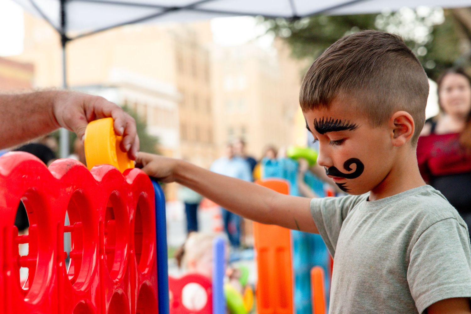 Young boy with mustache face paint plays a red Connect Four game at an outdoor festival.