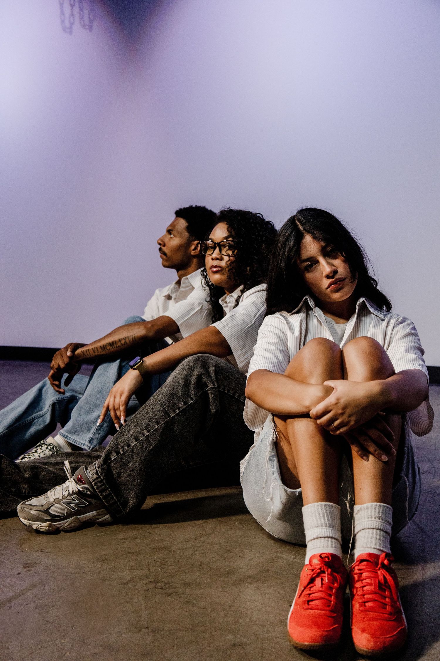 Three young adults in white shirts sitting against a wall in a moody studio setting.