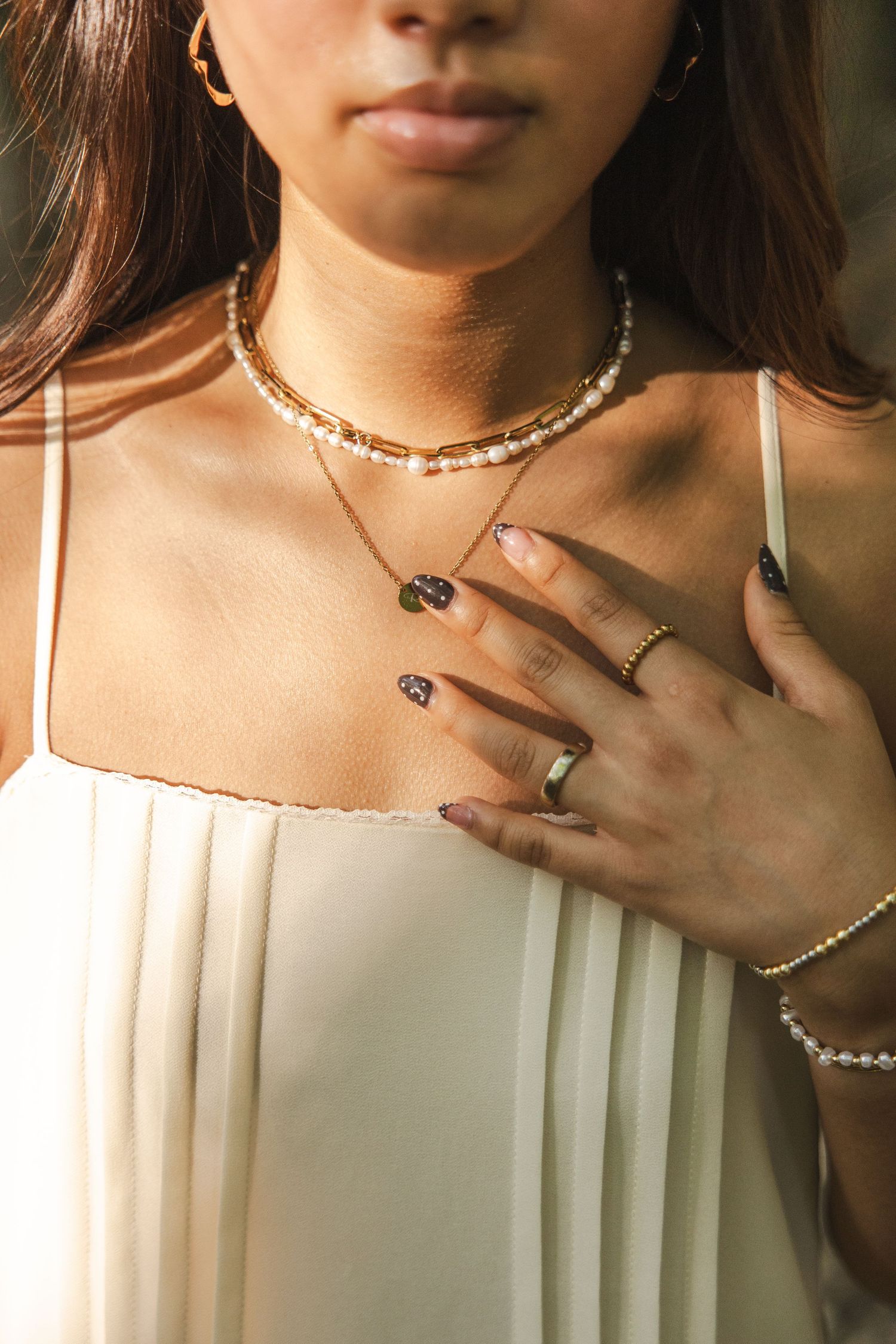Close-up of woman wearing layered gold and pearl necklaces with rings and bracelet in white dress.