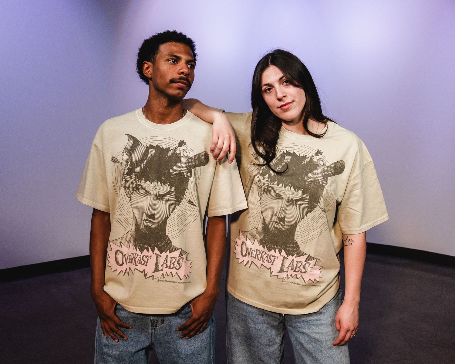 Two models wearing matching beige Outcast Labs graphic tees pose together in photo studio.