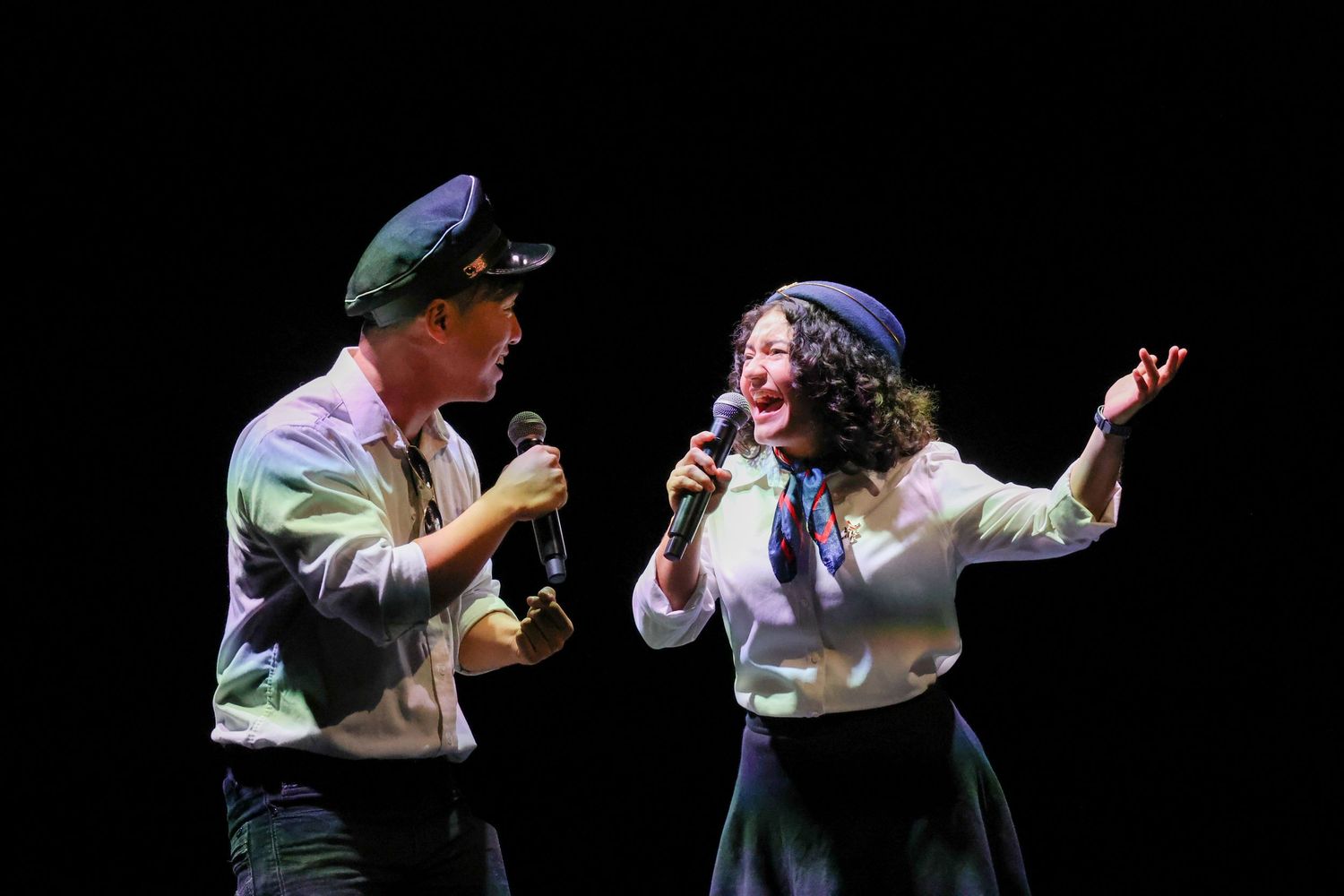 Two performers sing on a dark stage, one wearing a cap, during a theatrical cultural showcase.